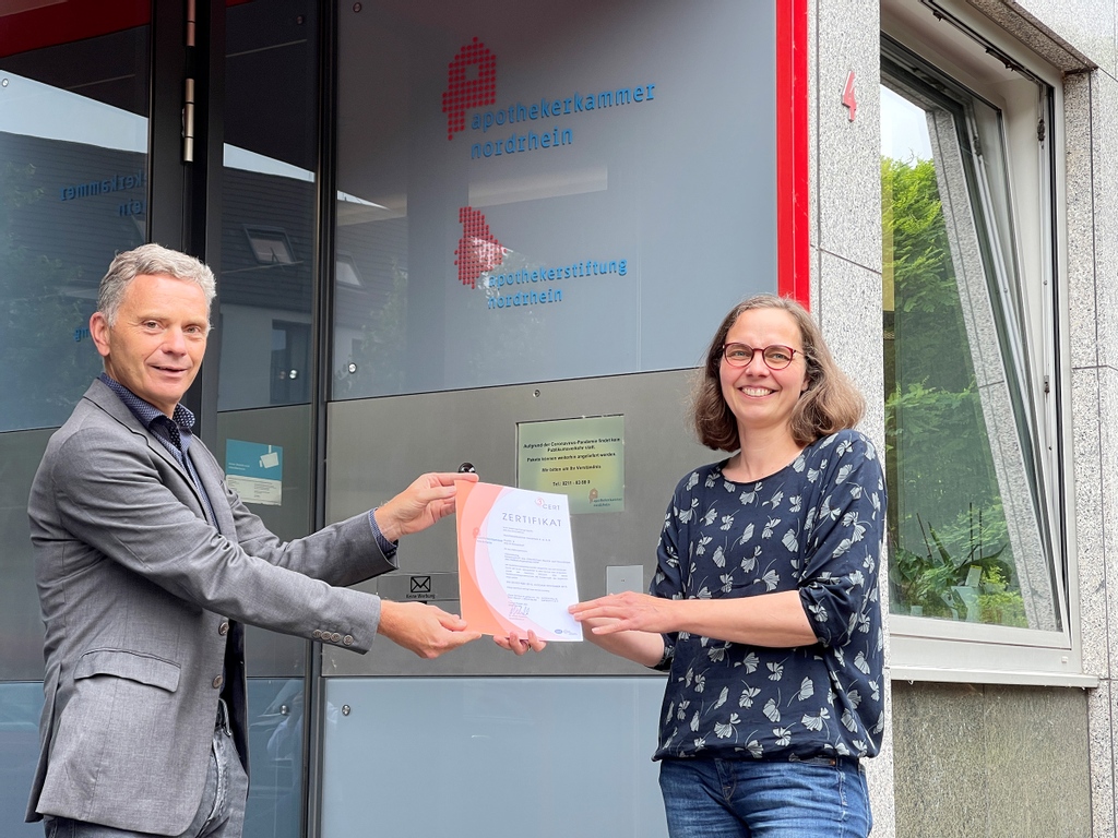 Auf diesem Bild erkennt man, wie der Geschäftsführer der Apothekerkammer Nordrhein Dr. Stefan Derix zusammen mit der QMS-Beauftragten Anne Tippel das QMS-Zertifikat der Apothekerkammer Nordrhein hält. Im Hintergrund ist der Eingang zur Geschäftsstelle zu sehen.