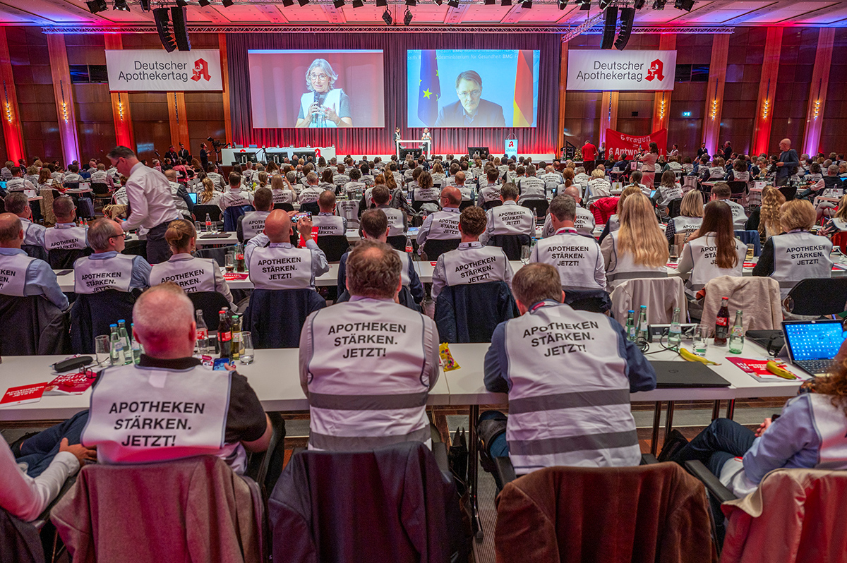 Delegierte des Deutschen Apothekertages sitzen im Plenum. Auf Leinwänden sind ABDA-Präsidentin Gabriele Regina Overwiening und Bundesgesundheitsminister Lauterbach zu sehen