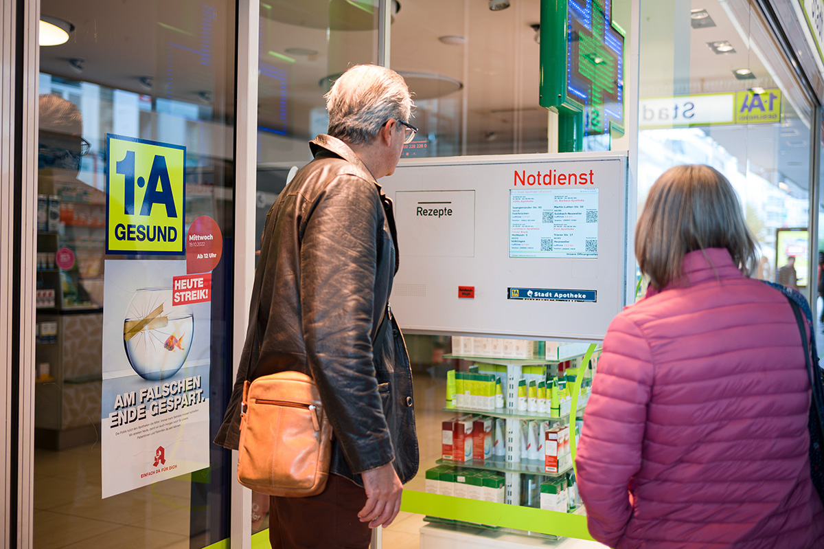 Patienten stehen vor einer geschlossenen Apotheke und schauen auf einen Aushang mit Notdienst-Apotheken