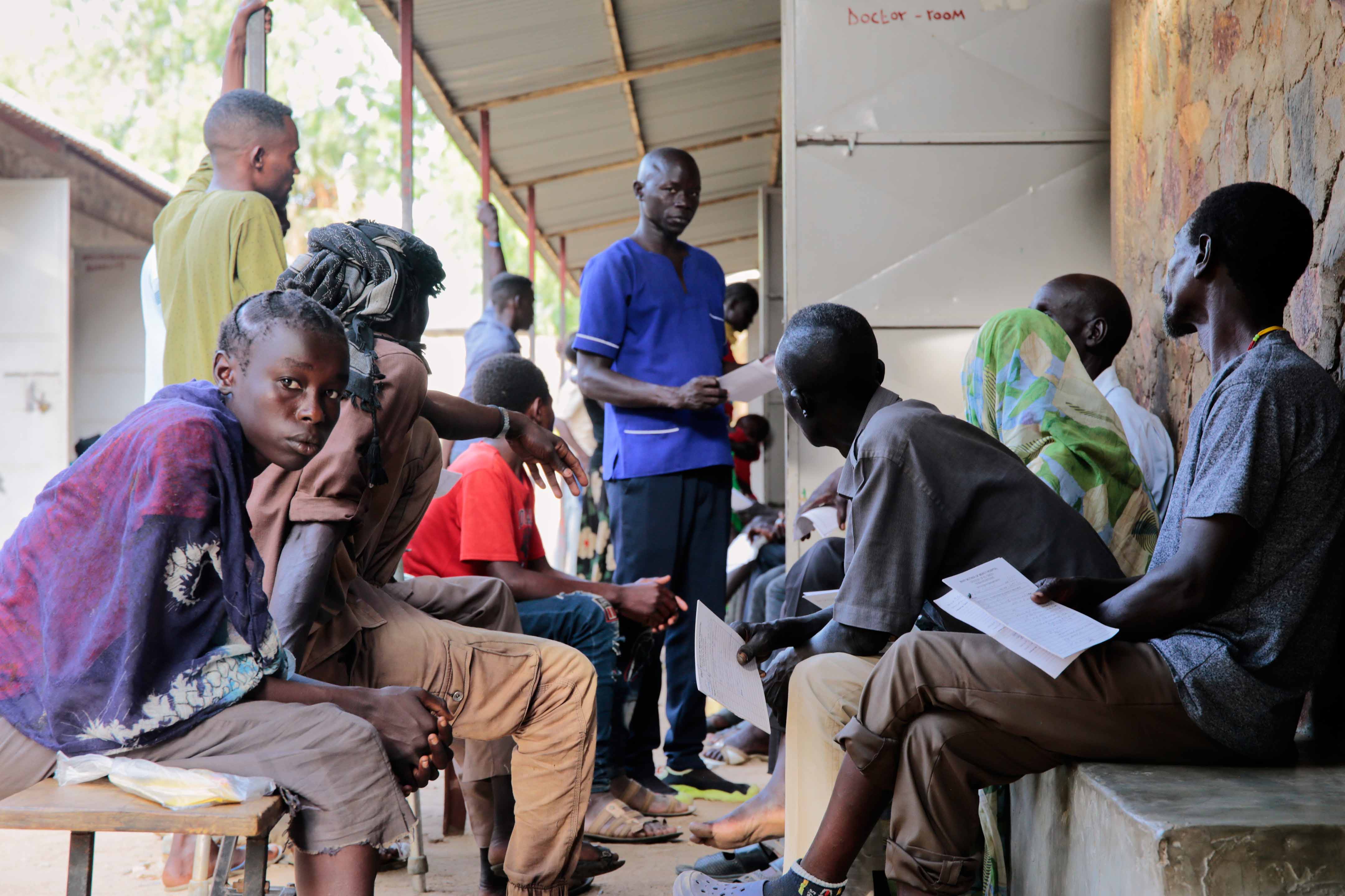 Menschen sitzen und stehen in einem überdachten Wartebereich, einige halten Dokumente in der Hand. Eine Person in blauer Kleidung steht in der Mitte und scheint Informationen oder Anweisungen zu geben.
