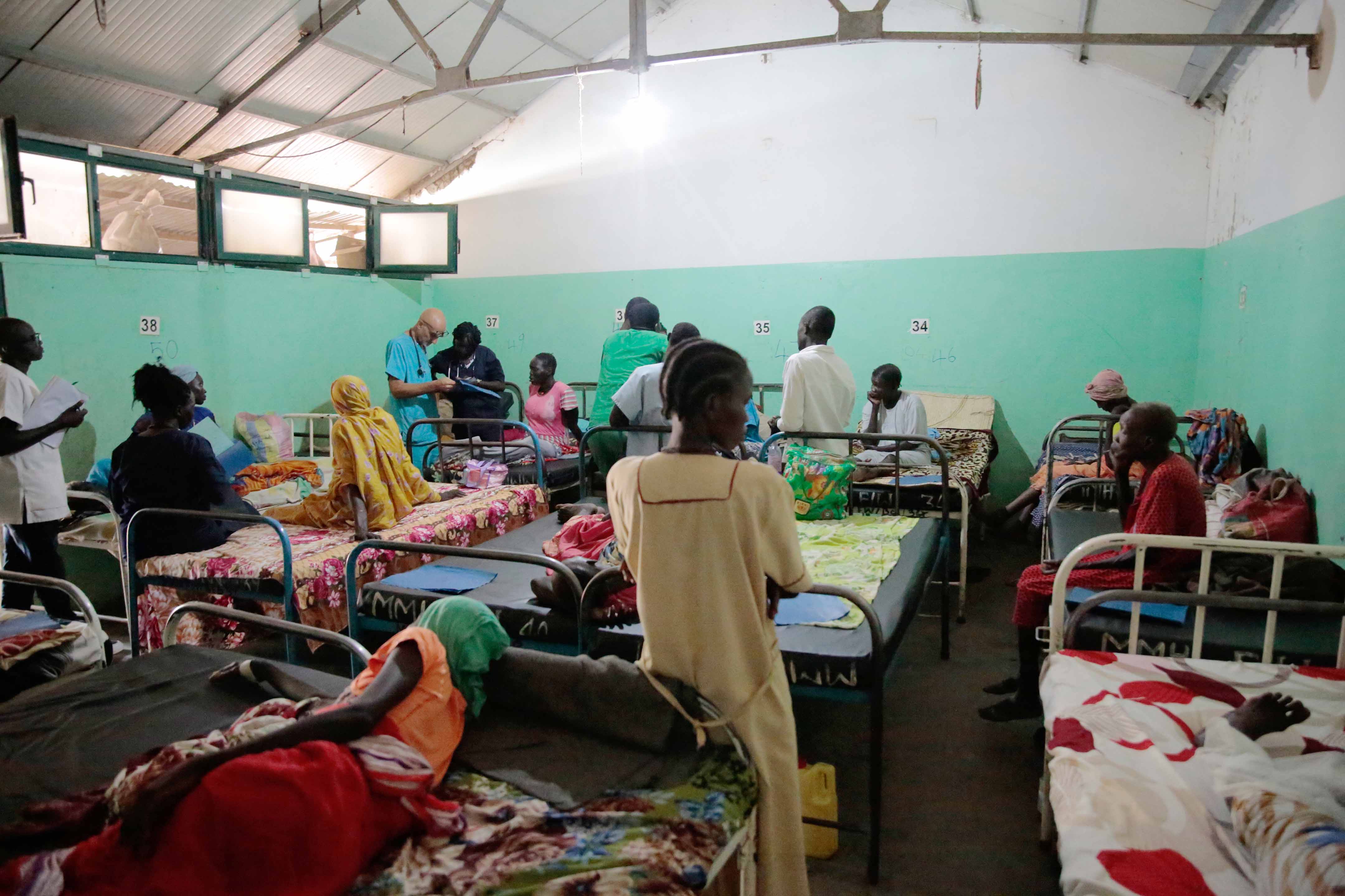 Krankenstation mit mehreren Betten, in denen Menschen liegen oder sitzen. Weitere Personen stehen oder gehen zwischen den Betten, einige halten Unterlagen. Die Wände sind grün gestrichen, und die Betten sind mit bunten Decken bedeckt.
