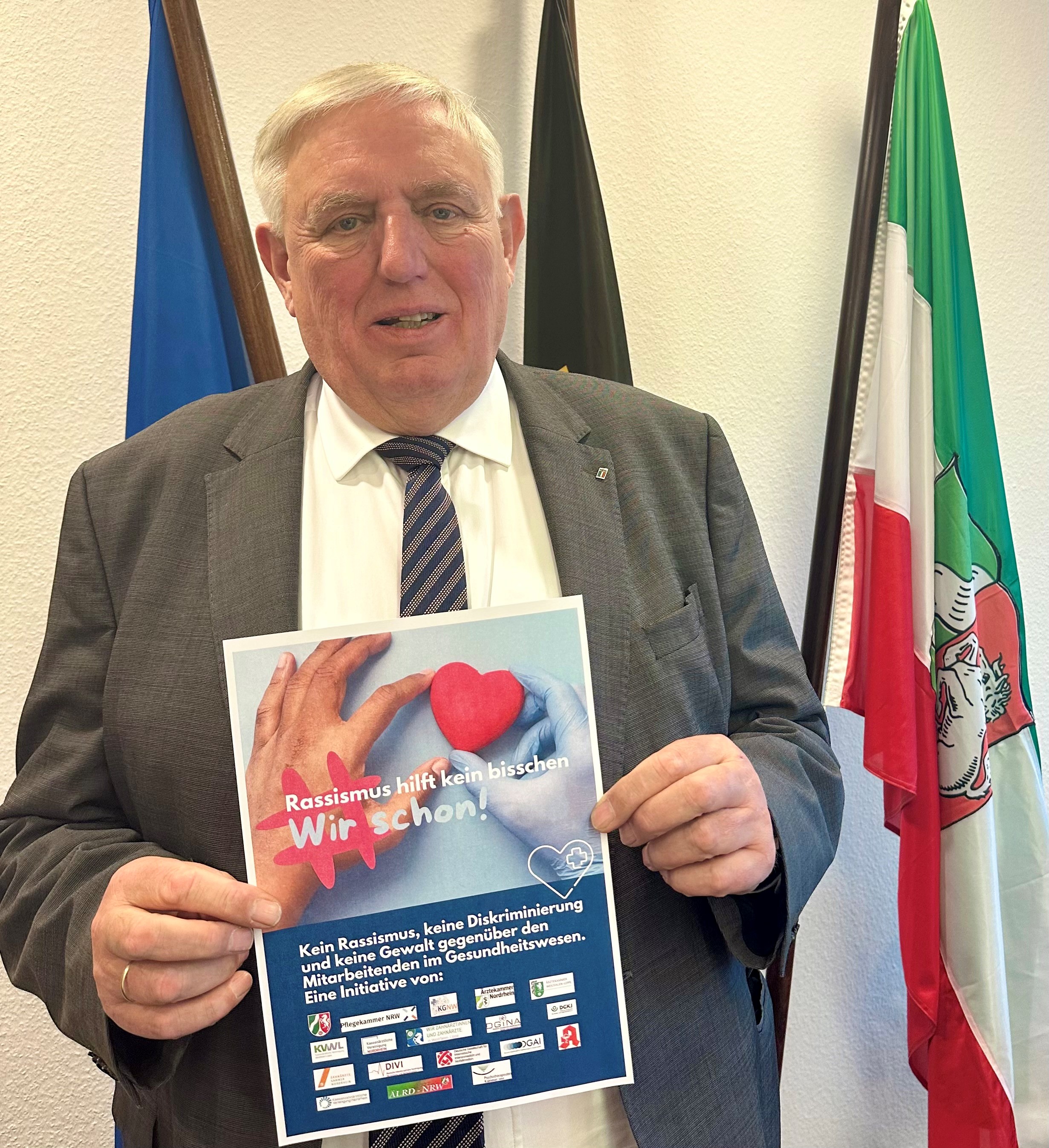 Gesundheitsminister Karl-Josef Lauman hällt einen ausdruck mit beiden Händen fest. Auf dem Ausdruck sind zwei Hände abgebildet, die zusammen ein Herz halten. Über dem Bild steht der Schriftzug "#Rassismus hilft kein bisschen Wir schon!" unter dem Bild sind alle Logos der an der Initiative beteiligten Organisationen.
