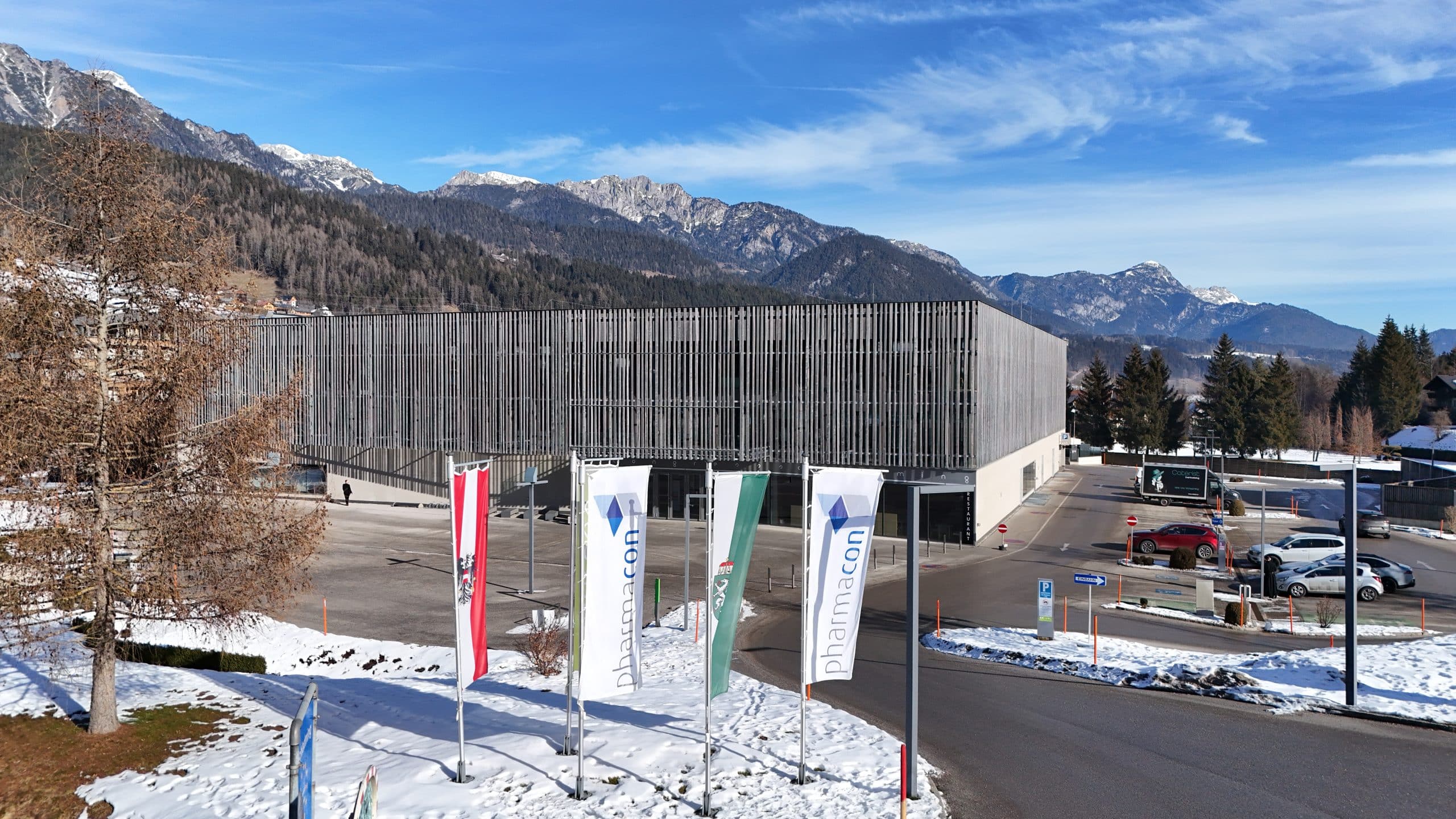 Modernes Kongressgebäude in winterlicher Berglandschaft mit schneebedecktem Boden und klar blauem Himmel. Vor dem Gebäude stehen mehrere Fahnen, darunter eine mit der Aufschrift ‚Pharmacon‘. Im Hintergrund sind Berge und Bäume zu sehen.