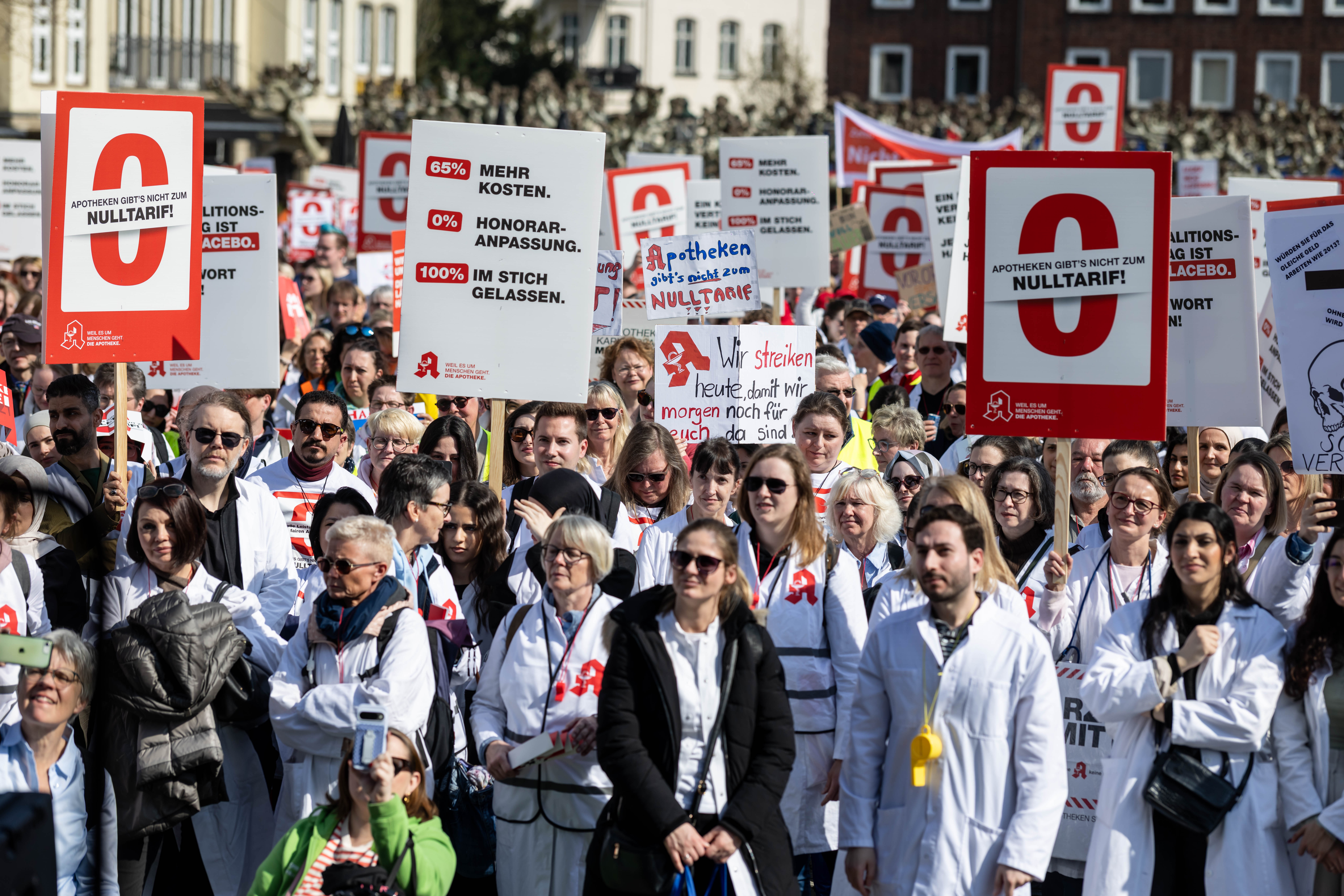 Eine große Gruppe von Menschen in weißen Arbeitskitteln steht dicht gedrängt bei einer öffentlichen Demonstration. Viele Personen halten Plakate mit roten und weißen Gestaltungselementen hoch. Die Schilder tragen Texte wie „0 % Honorar-Anpassung“, „Apotheken gibt’s nicht zum Nulltarif“ und „85 % mehr Kosten“. Im Hintergrund sind Gebäude und weitere Teilnehmende zu sehen. Die Szene spielt im Freien bei sonnigem Wetter.