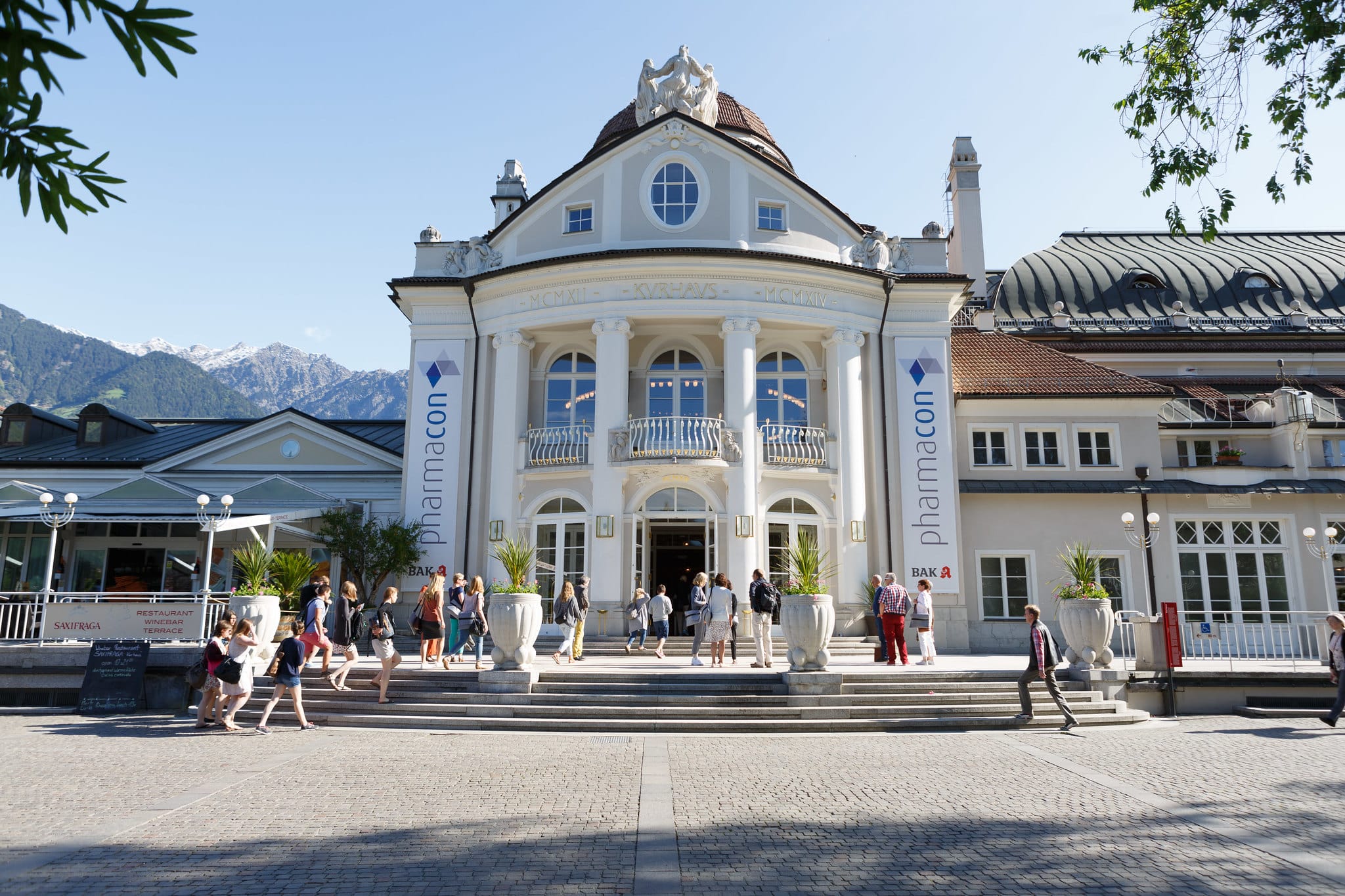 Historisches Kurhaus Meran mit klassizistischer Fassade und Säulen, davor Menschen auf dem Vorplatz; an den Säulen hängen Banner mit der Aufschrift ‚pharmacon‘, im Hintergrund Berglandschaft bei sonnigem Wetter
