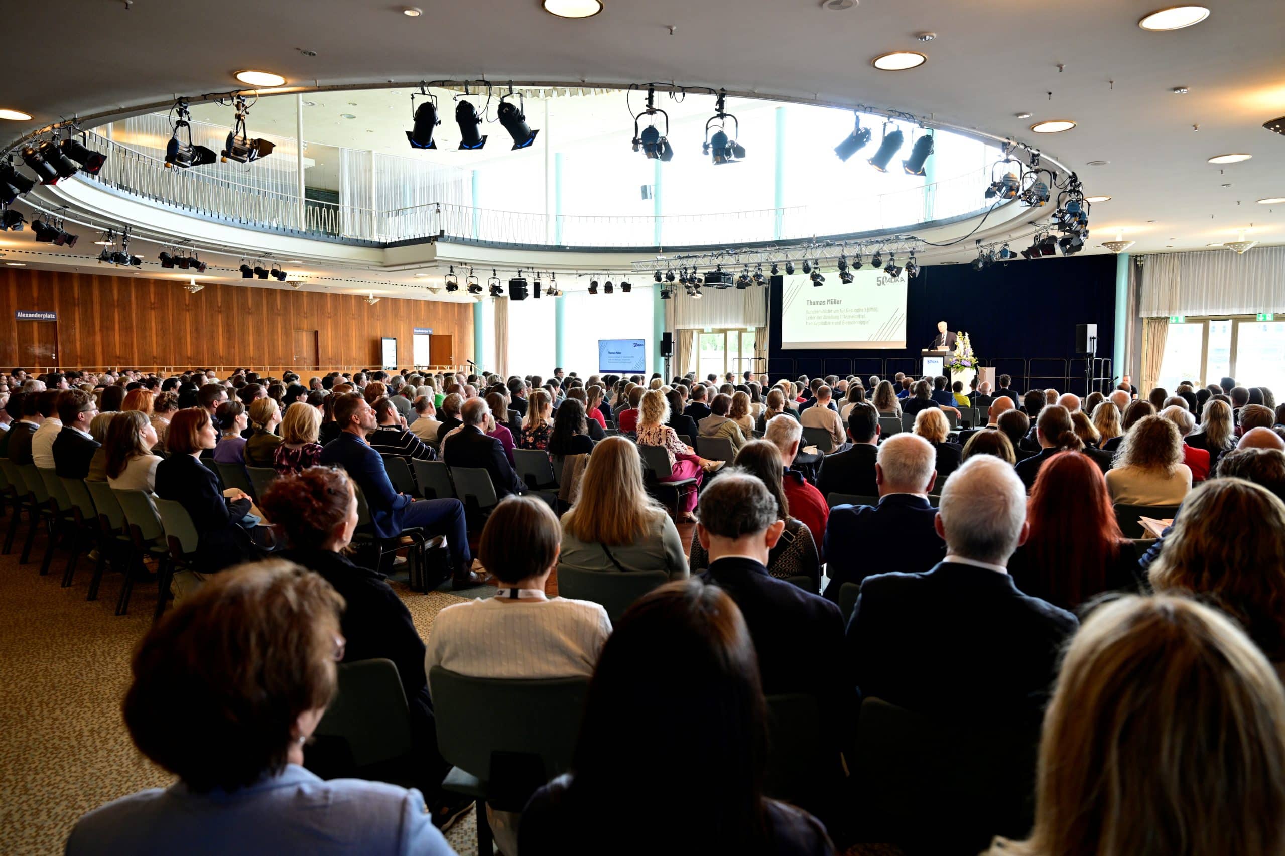 Großer Vortragssaal mit hunderten Teilnehmerinnen und Teilnehmern, die einem Vortrag auf einer Bühne folgen. Die helle, moderne Veranstaltungsstätte ist gut gefüllt, mit Blick auf die Präsentationsfläche und große Fenster im Hintergrund.