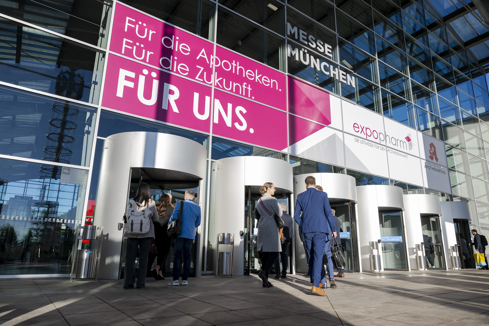 Menschen betreten das moderne Glasgebäude der Messe München durch Drehkreutztüren, während über dem Eingang große rosa und weiße Banner mit der Aufschrift „Für die Apotheken. Für die Zukunft. FÜR UNS.“ sowie Hinweise auf die Messe „expopharm“ hängen.
