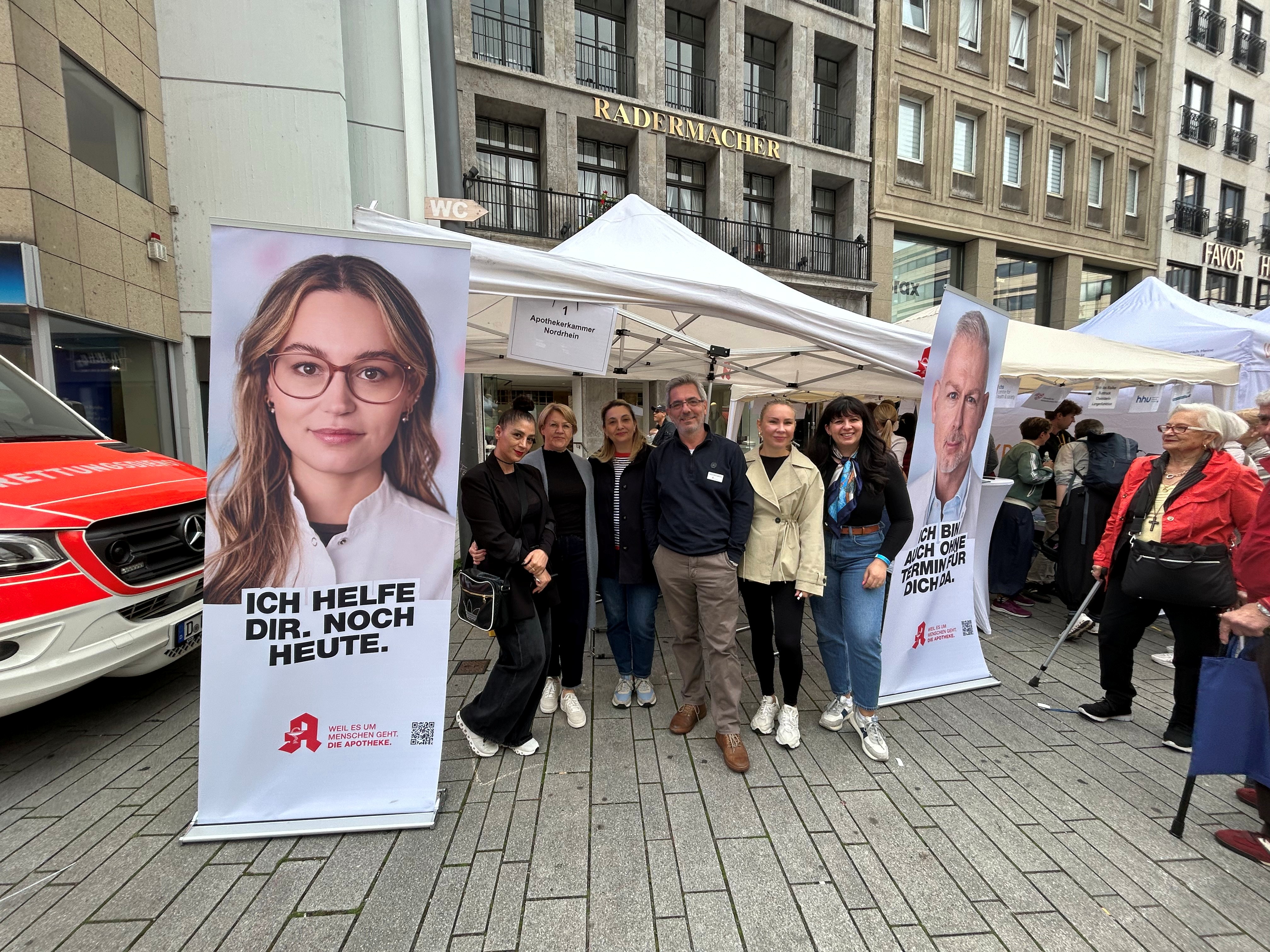 Stand der Apothekerkammer Nordrhein beim Gesundheitstag auf dem Düsseldorfer Schadowplatz: Mehrere Teammitglieder stehen vor einem Informationszelt zwischen zwei Roll-Ups der Apothekenkampagne. Besucherinnen und Besucher informieren sich am Stand über Gesundheitsvorsorge, die elektronische Patientenakte und Berufe in der Apotheke. Im Hintergrund sind weitere Aktionsstände sowie ein Rettungswagen zu sehen.