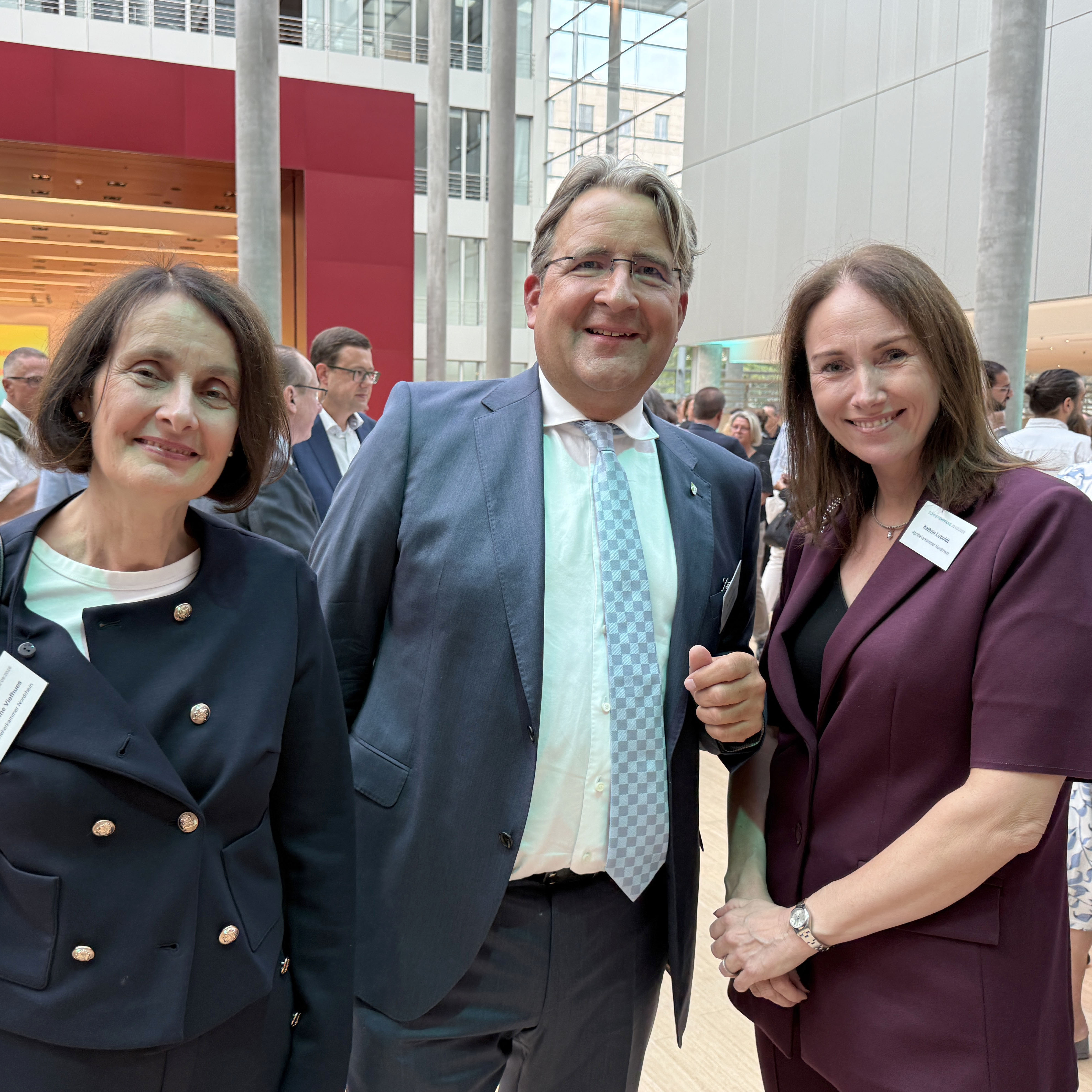 Drei Personen stehen bei einem Empfang in einem modernen, lichtdurchfluteten Atrium mit hohen Glasflächen, Säulen und einer roten Wand im Hintergrund. Die Personen tragen Business-Kleidung und Namensschilder und posieren für die Kamera. Im Hintergrund sind weitere Gäste der Veranstaltung zu sehen, die sich in kleinen Gruppen unterhalten. Die Aufnahme vermittelt den Eindruck einer Netzwerk- oder Abendveranstaltung in repräsentativem Ambiente.