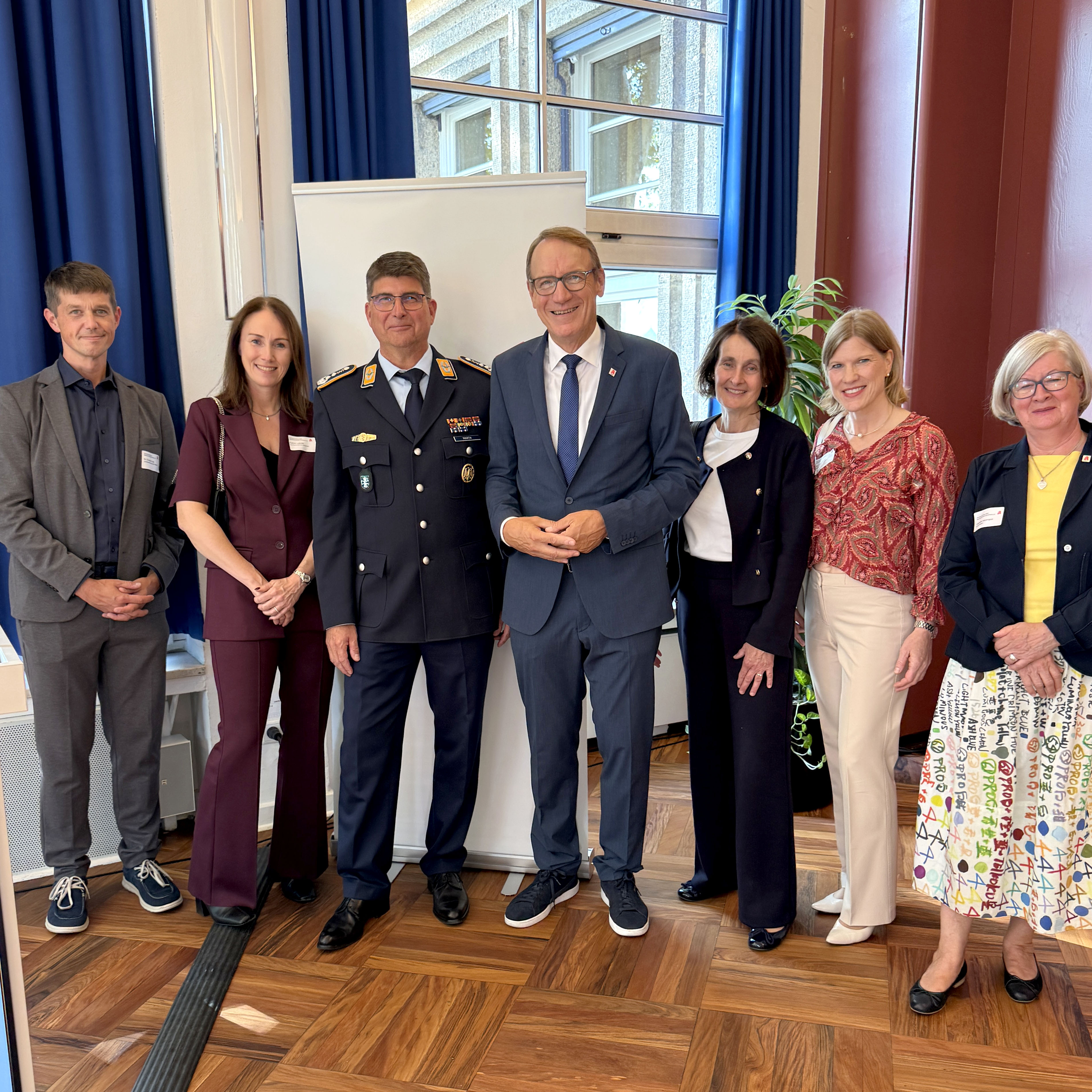 Sieben Personen stehen für ein Gruppenfoto in einem hellen Veranstaltungsraum mit hohen Fenstern, blauen Vorhängen und Parkettboden. In der Mitte stehen zwei Personen, eine davon in dunkler Uniform, umgeben von fünf weiteren Personen in Business-Kleidung. Im Hintergrund sind ein Roll-up-Aufsteller sowie eine Zimmerpflanze zu sehen. Die Gruppe blickt zur Kamera.