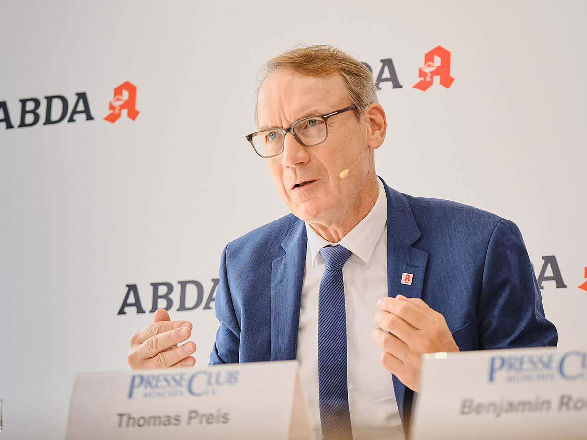 Person in blauem Anzug mit Brille sitzt an einem Podiumstisch vor einer ABDA-Pressewand und spricht während einer Pressekonferenz. Im Vordergrund ist ein Namensschild sichtbar, im Hintergrund sind mehrfach das ABDA-Logo und der Veranstaltungsort PresseClub München zu sehen.