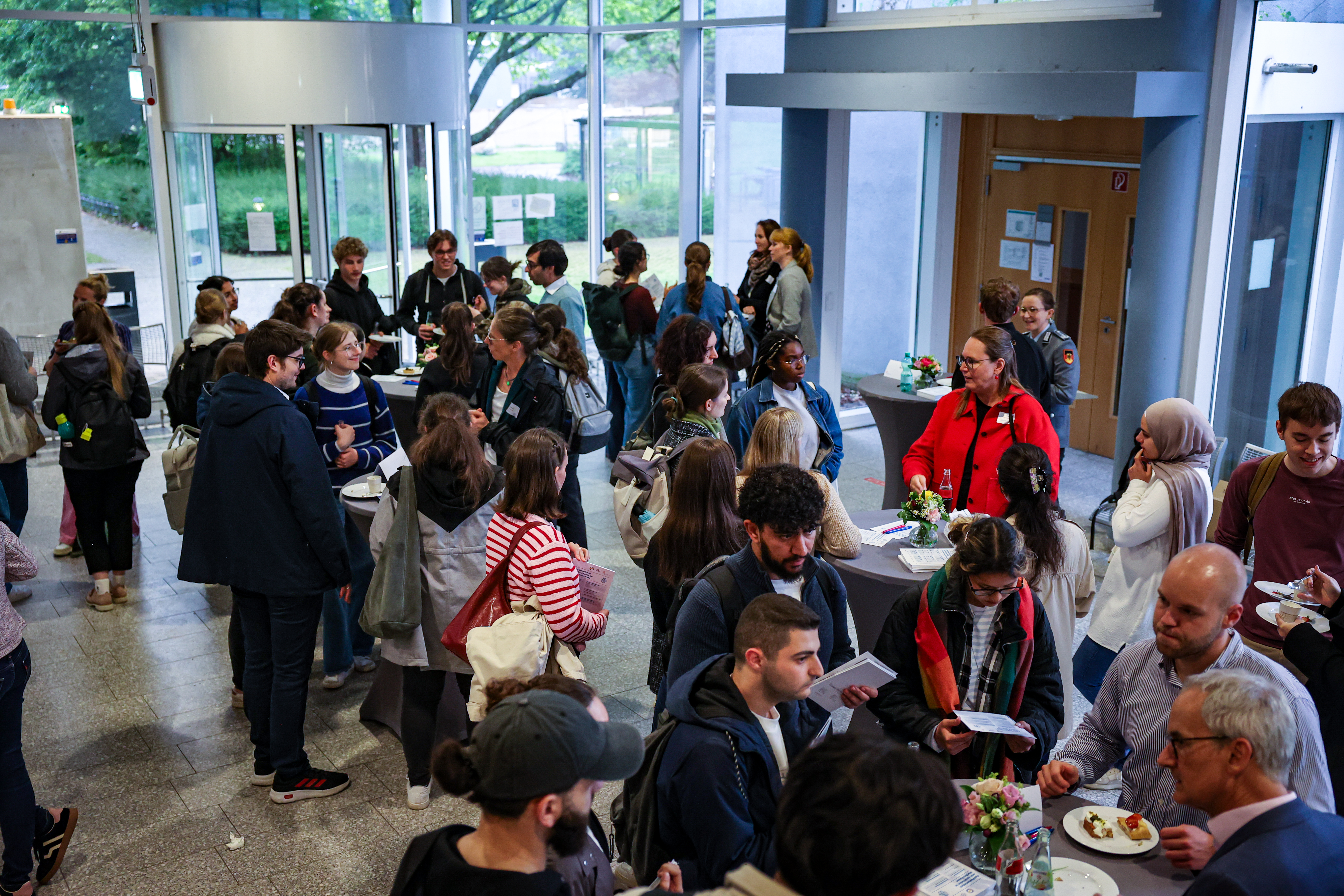 Große Gruppe von Studierenden und Referierenden steht in einem hellen Foyer und tauscht sich in kleinen Gruppen aus. Mehrere Stehtische mit Getränken, Snacks und Informationsmaterialien sind verteilt im Raum. Durch große Glasflächen im Hintergrund fällt Tageslicht, während im Vordergrund Personen miteinander ins Gespräch vertieft sind und Flyer in den Händen halten.