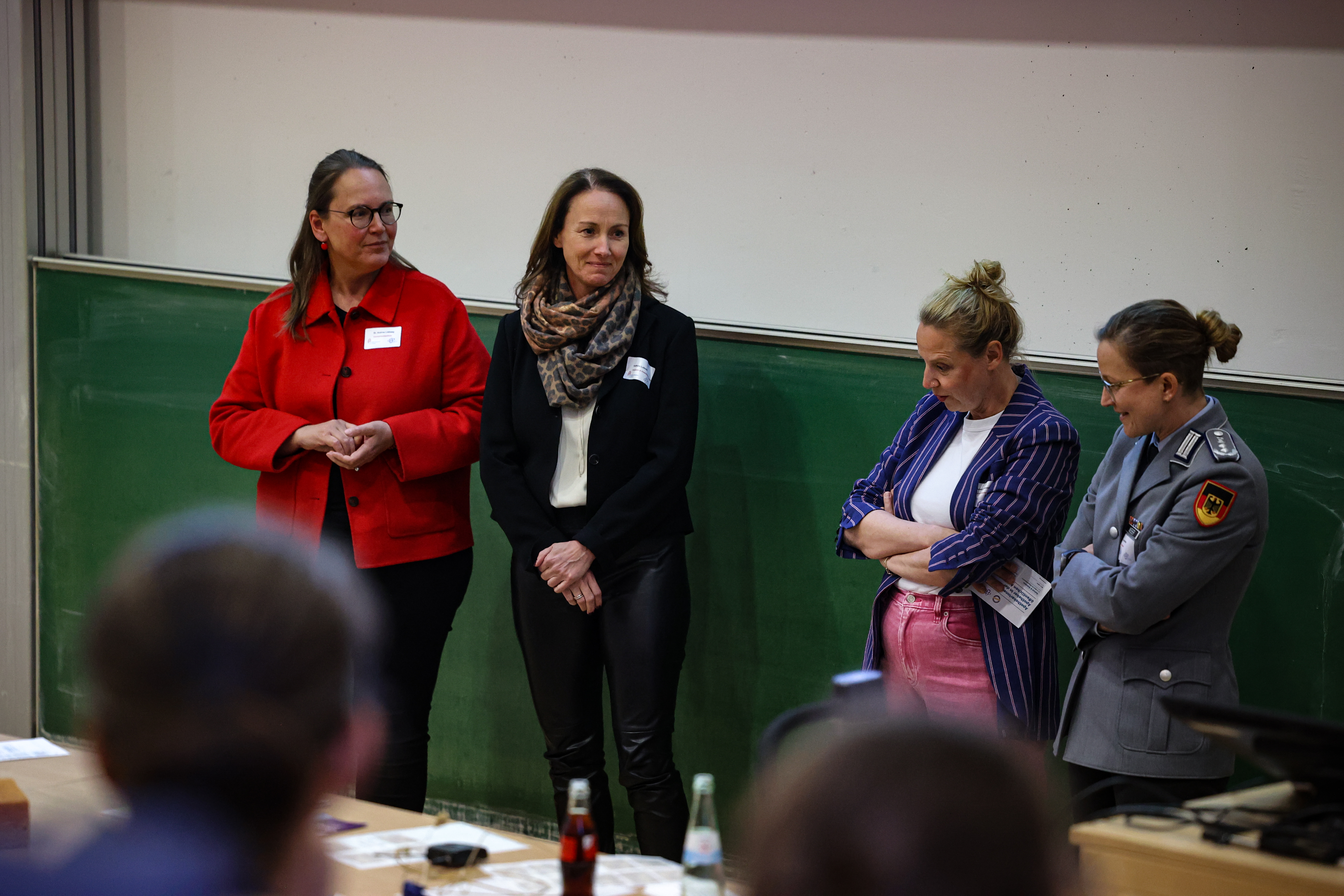 Vier Personen stehen vor einer Tafel in einem Hörsaal und nehmen an einer moderierten Diskussionsrunde teil. Die Personen tragen Namensschilder und sind unterschiedlich gekleidet, darunter ein roter Mantel, ein dunkles Outfit mit Schal, ein gestreiftes Sakko sowie eine Uniform. Im Vordergrund sind unscharf sitzende Zuhörende sowie Tische mit Getränkeflaschen zu erkennen.