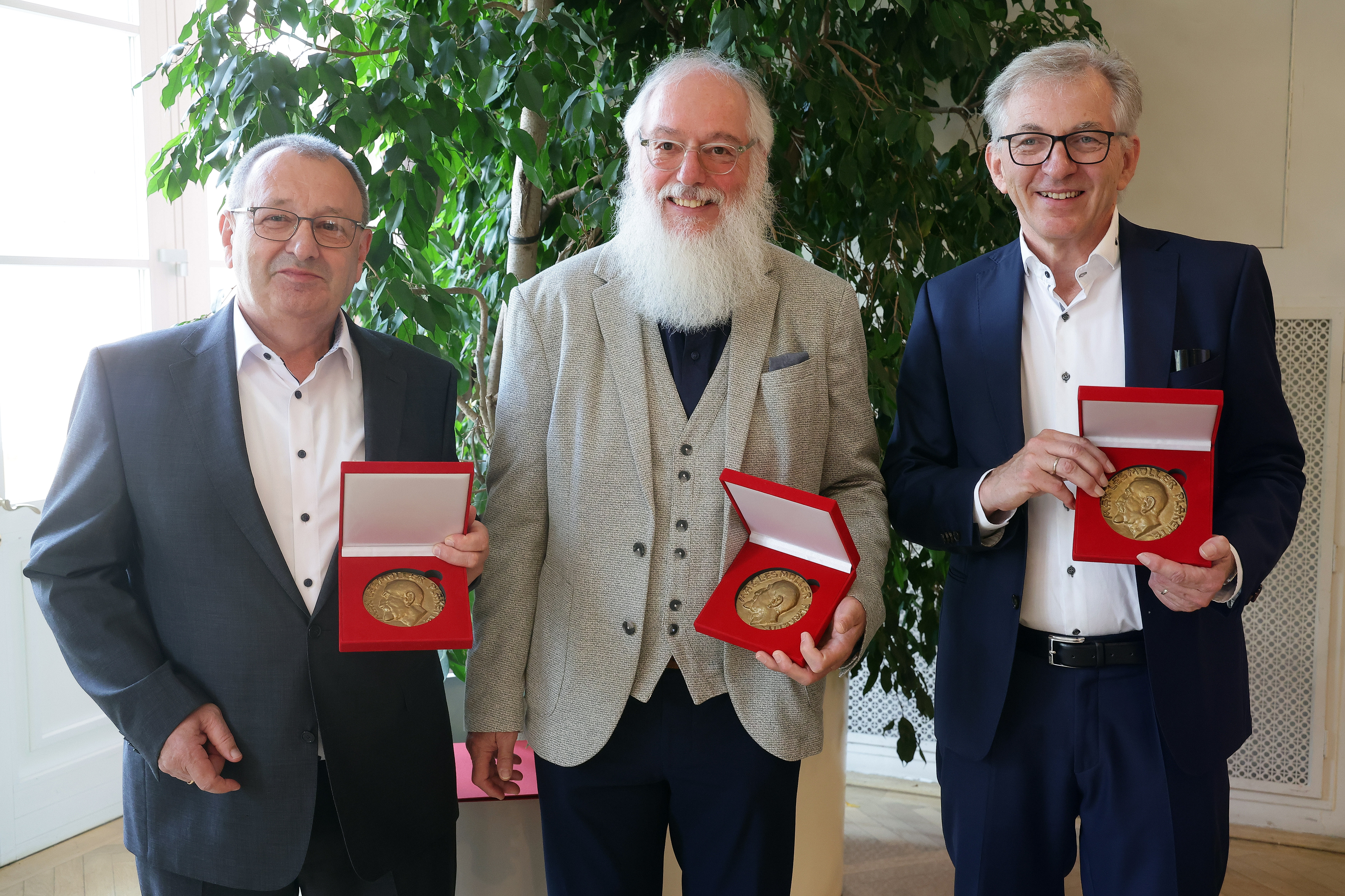 Drei Personen stehen nebeneinander in einem Innenraum vor einer großen Pflanze und halten jeweils eine geöffnete rote Schmuckschatulle mit einer goldenen Medaille in der Hand. Die Medaillen sind gut sichtbar präsentiert; es handelt sich um die Lesmüller-Medaille, mit der die drei Ausgezeichneten geehrt wurden.