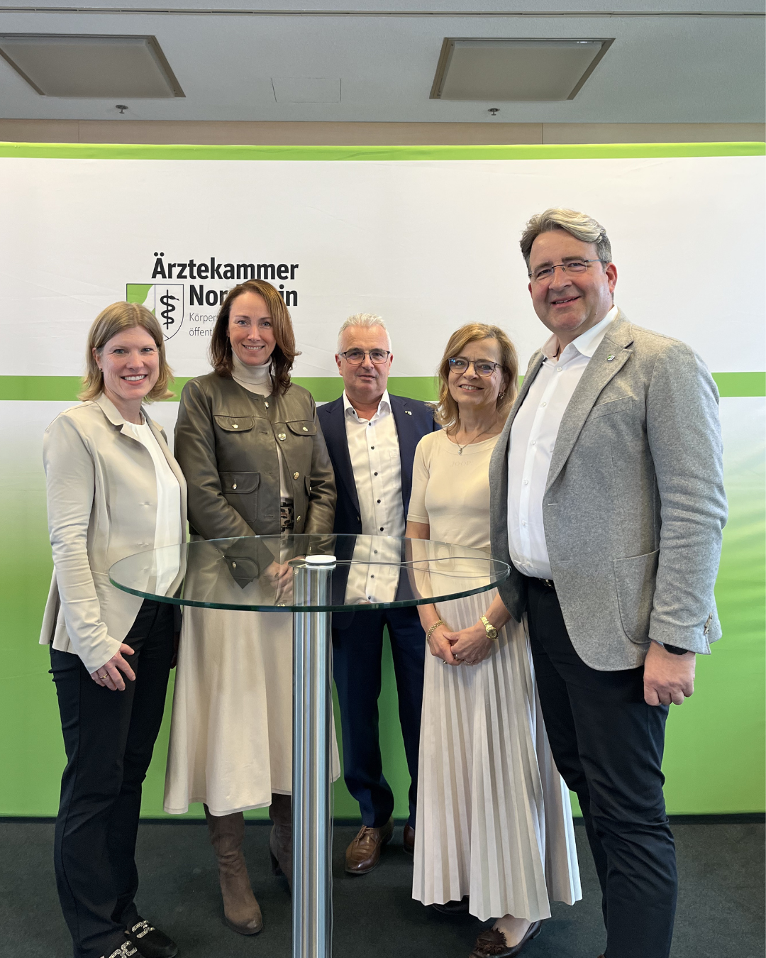 ünf Personen stehen lächelnd vor einem Hintergrund mit dem Logo der Ärztekammer Nordrhein. Sie posieren um einen runden Glastisch.