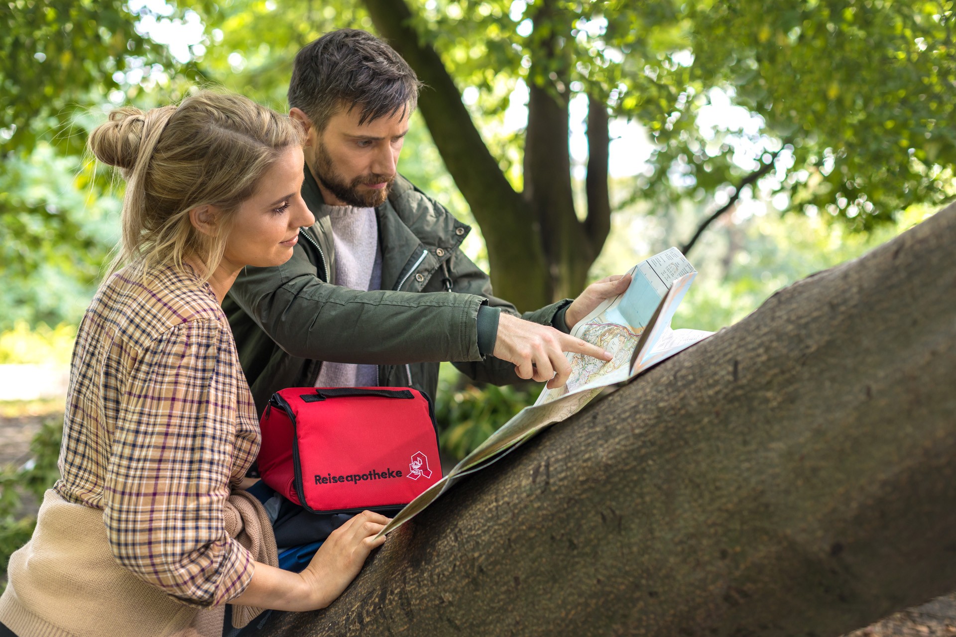 Zwei Personen orientieren sich mit einer aufgefalteten Landkarte in einer grünen Park- oder Waldlandschaft. Auf einem Baumstamm liegt eine rote Tasche mit der Aufschrift „Reiseapotheke“ und dem Apotheken-A-Symbol. Die Personen betrachten gemeinsam die Karte, während eine Person auf einen Ort auf der Karte zeigt. Im Hintergrund sind Bäume und dichtes Laub zu sehen.