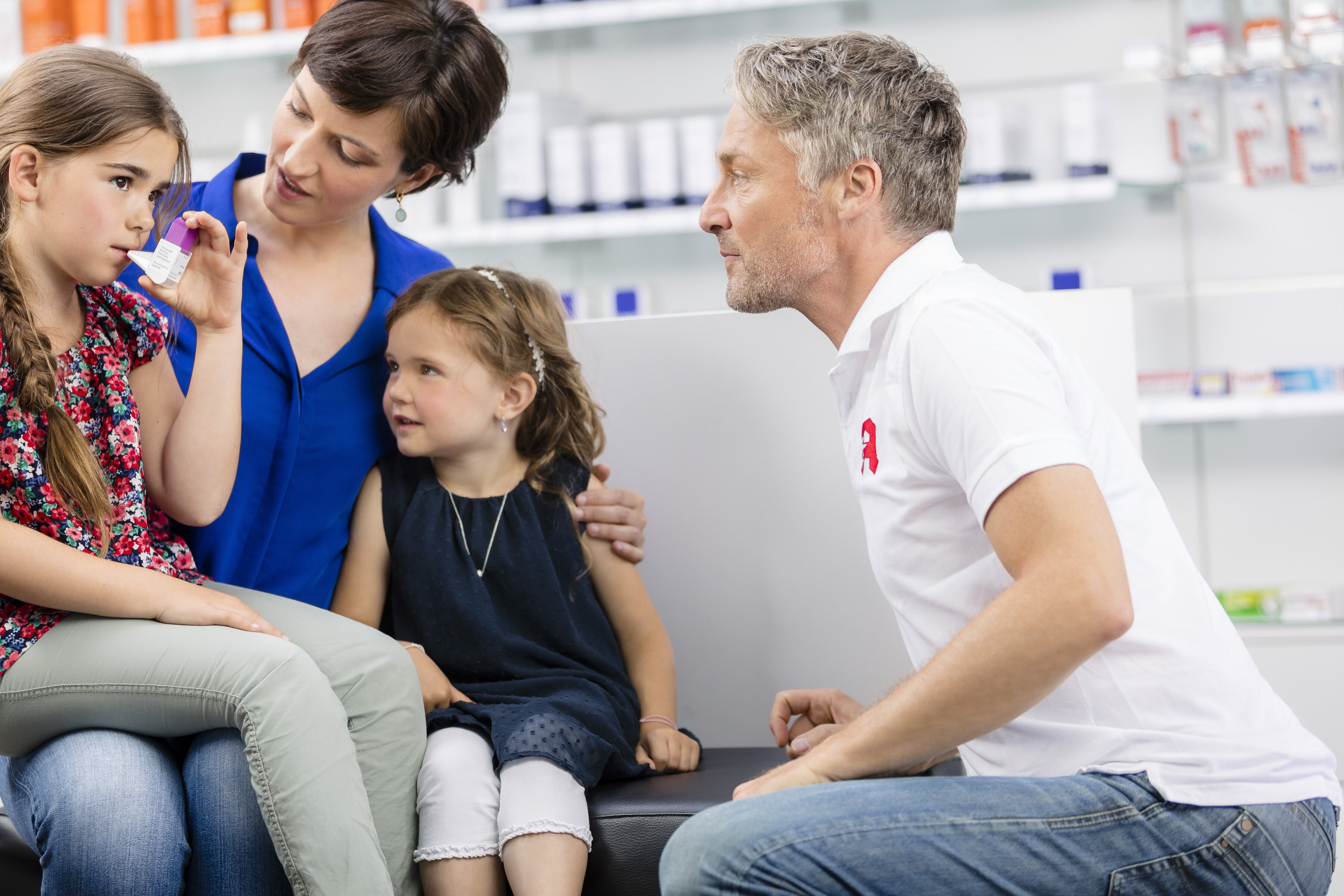 Ein Apotheker zeigt einem Kind die Anwendung eines Inhalators, während ein weiteres Kind daneben sitzt; eine weitere Person in blauer Kleidung beobachtet die Situation in einer hellen Apotheke mit Regalen im Hintergrund.