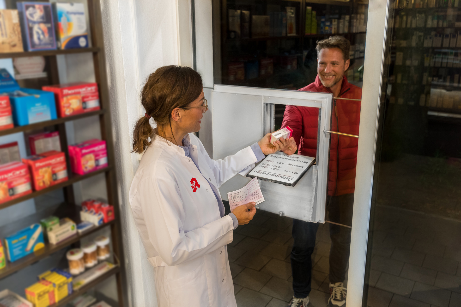 Eine Apothekerin reicht ein Medikament nachts durch die Notdienst-Klappe einer Apotheke an einen Patienten