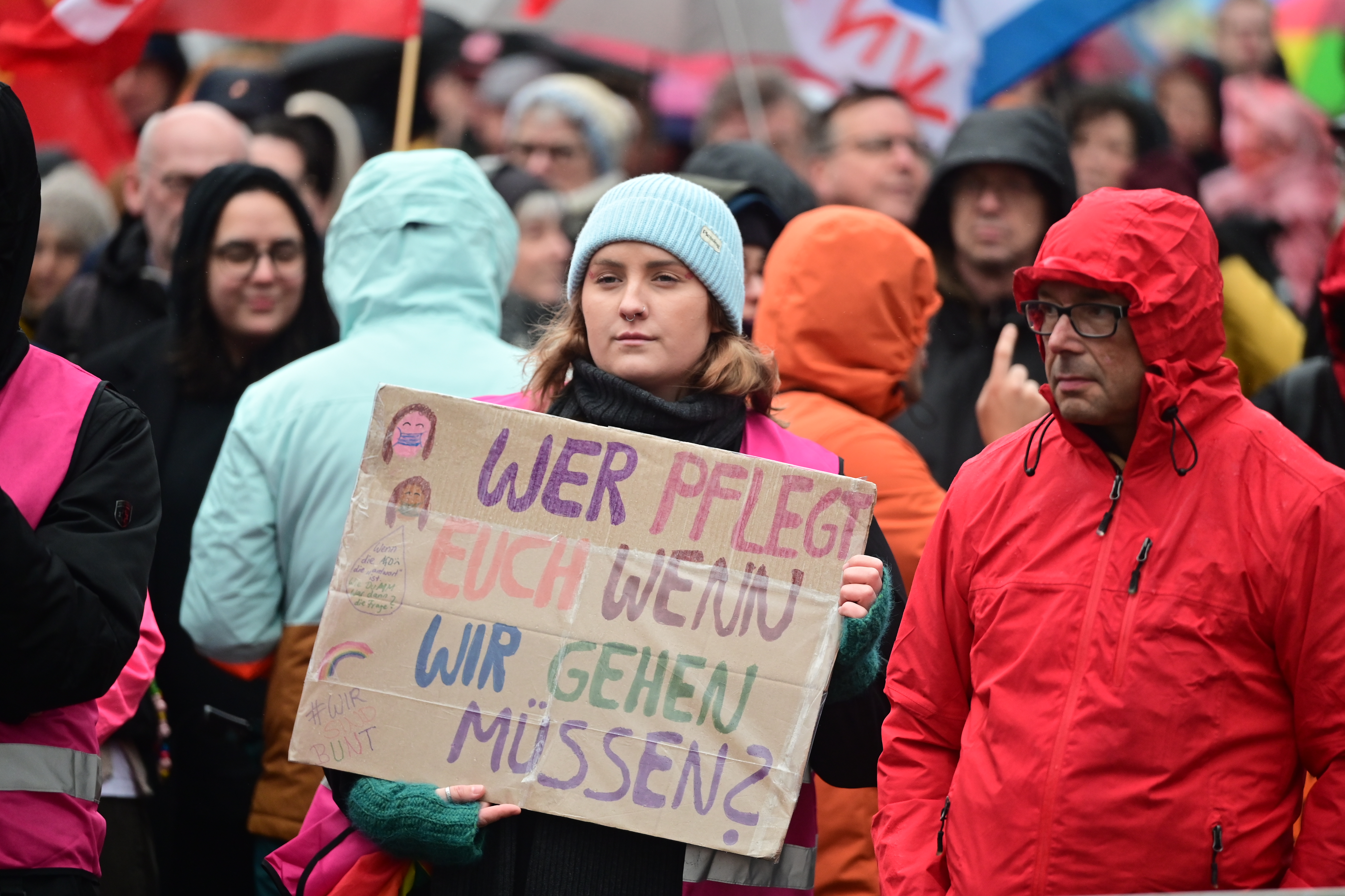 Eine Frau hält in Mitten einer Demonstration ein selbstgestaltetes Plakat in den Händen. Auf dem Plakat steht geschrieben: "WER PFLEGT EUCH WENN WIR GEHEN MÜSSEN?"