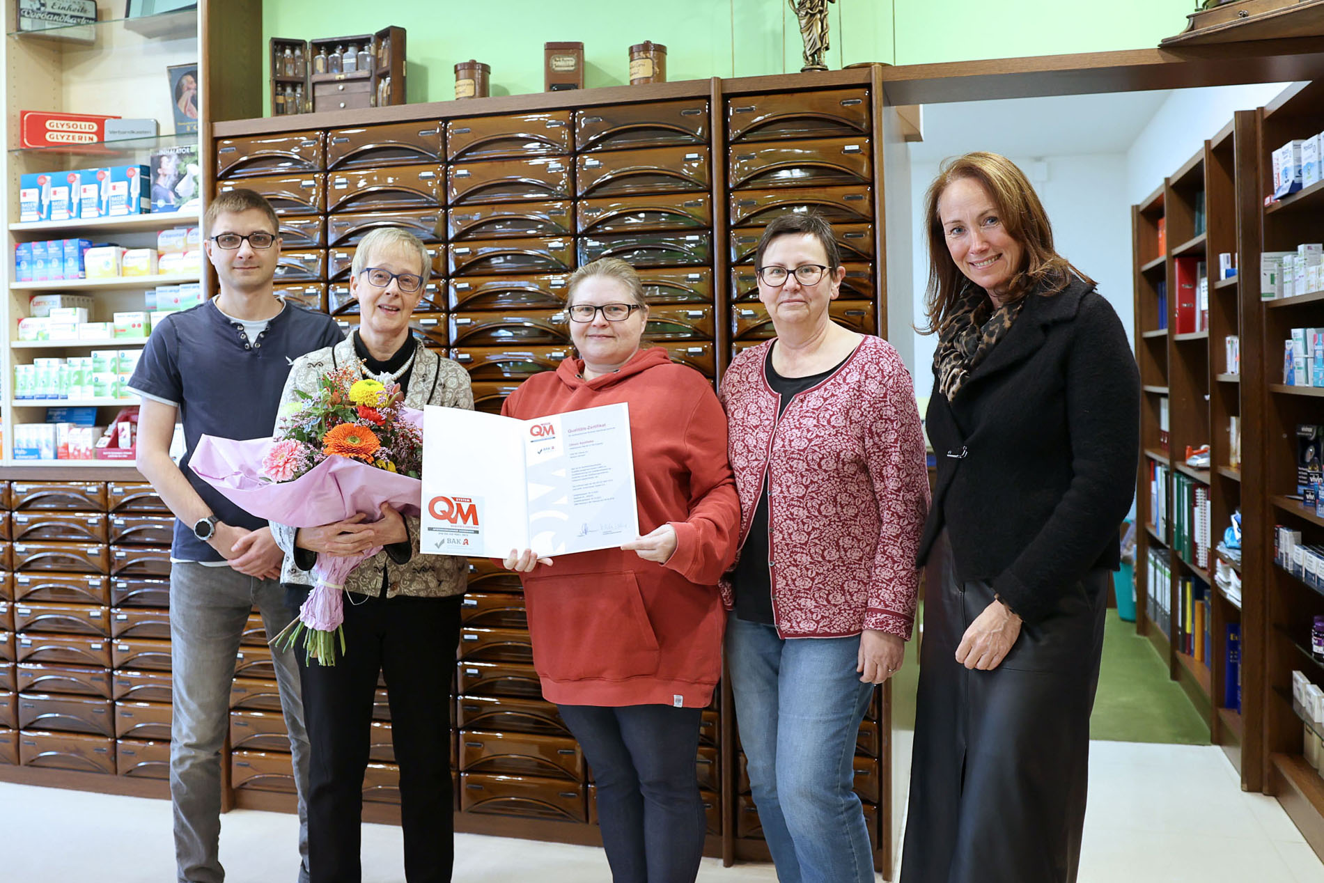 Gruppenfoto in einer Apotheke: Fünf Personen stehen vor einer Schubladenwand, eine Person hält einen Blumenstrauß, eine andere präsentiert ein offenes Zertifikat mit QM-Logo.