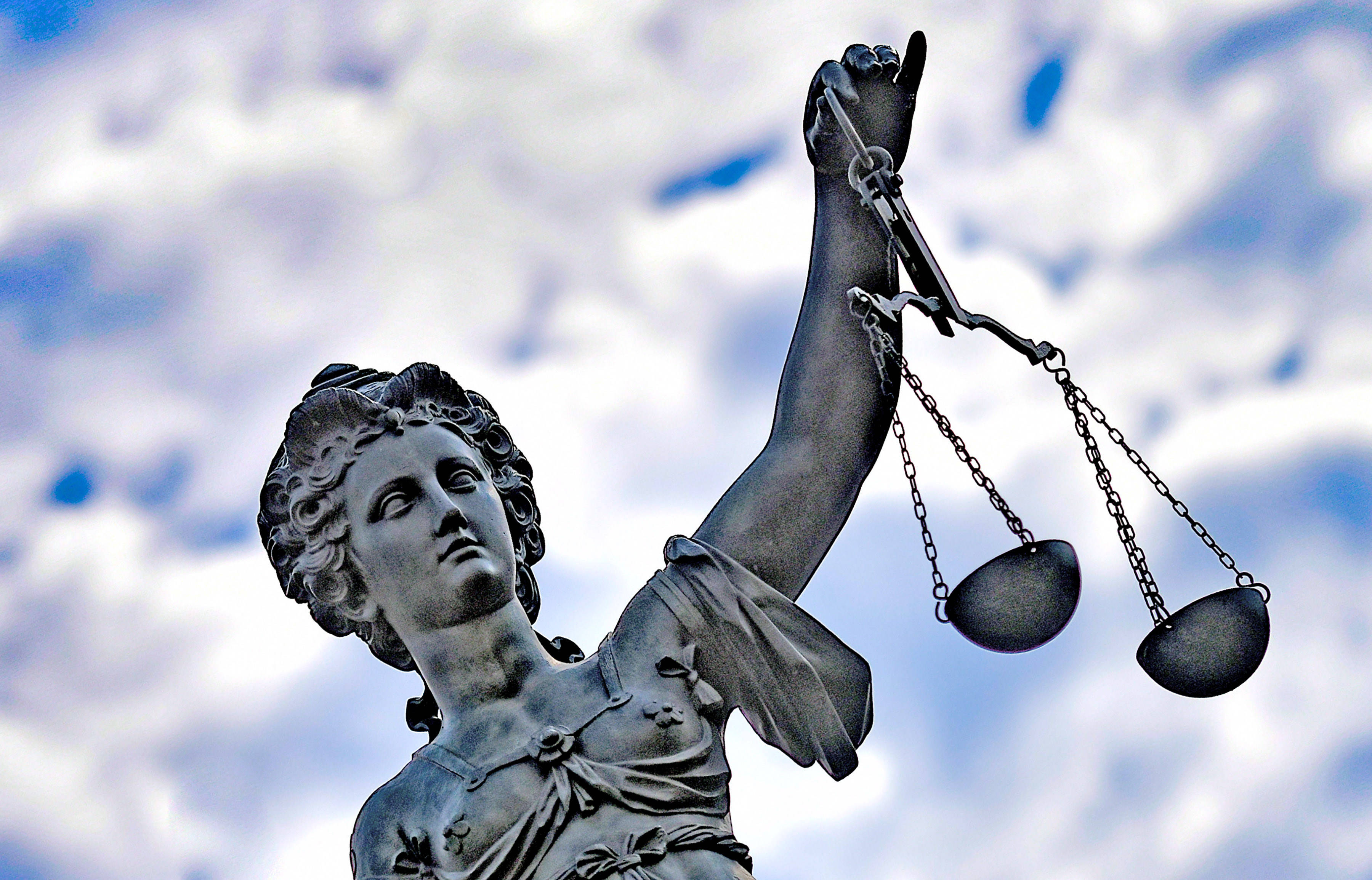 Statue der Justitia mit erhobenem Arm, in dem sie eine Waage hält. Die Skulptur ist aus dunklem Metall gefertigt und steht vor einem Himmel mit weißen Wolken.