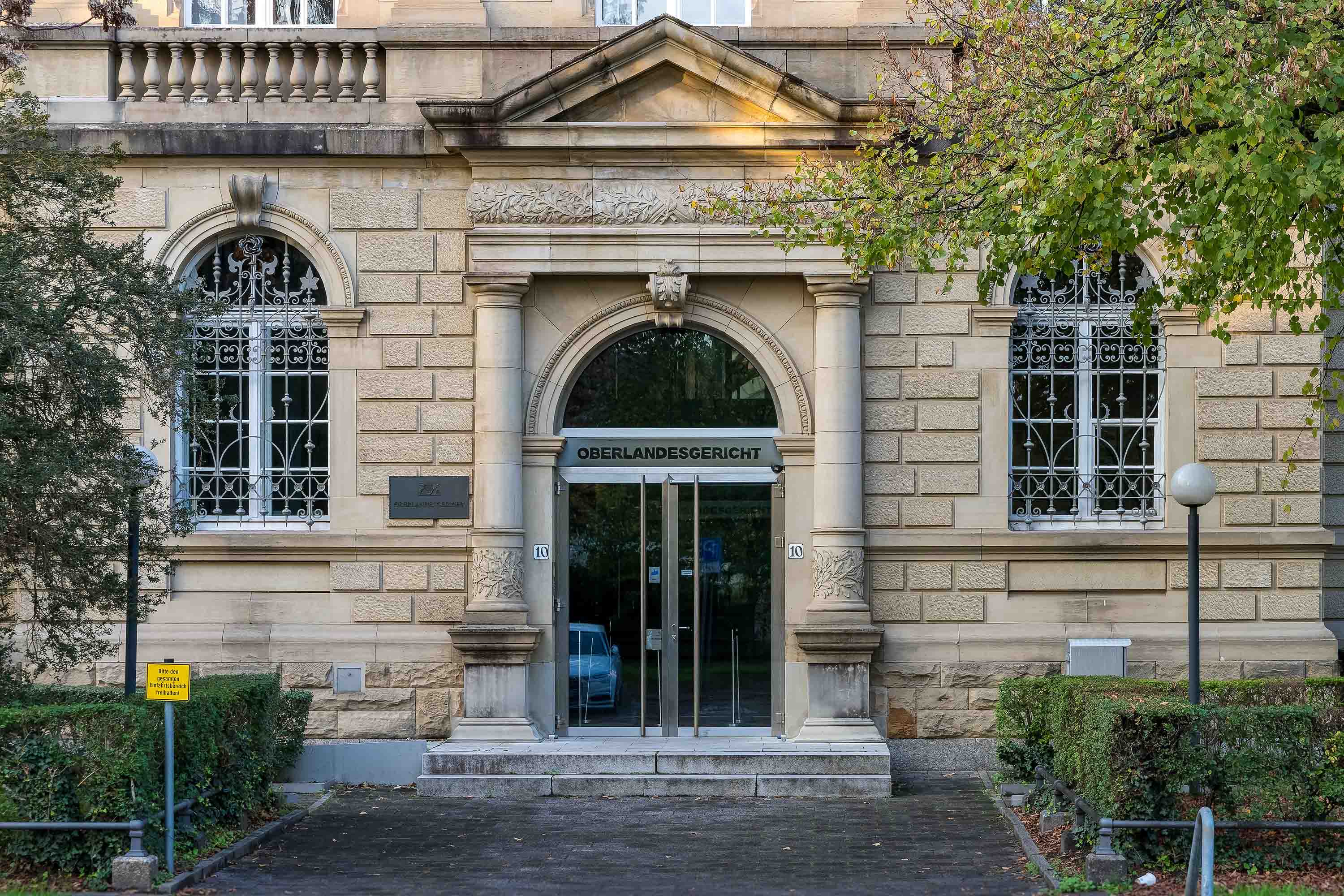 Eingang eines historischen Gebäudes mit der Aufschrift ‚Oberlandesgericht‘ über einer Glasdoppeltür. Die Fassade besteht aus hellem Sandstein mit dekorativen Elementen, links und rechts sind große Fenster mit kunstvollen Gittern. Vor dem Gebäude befinden sich ein gepflasterter Weg, Grünflächen und Laternen.
