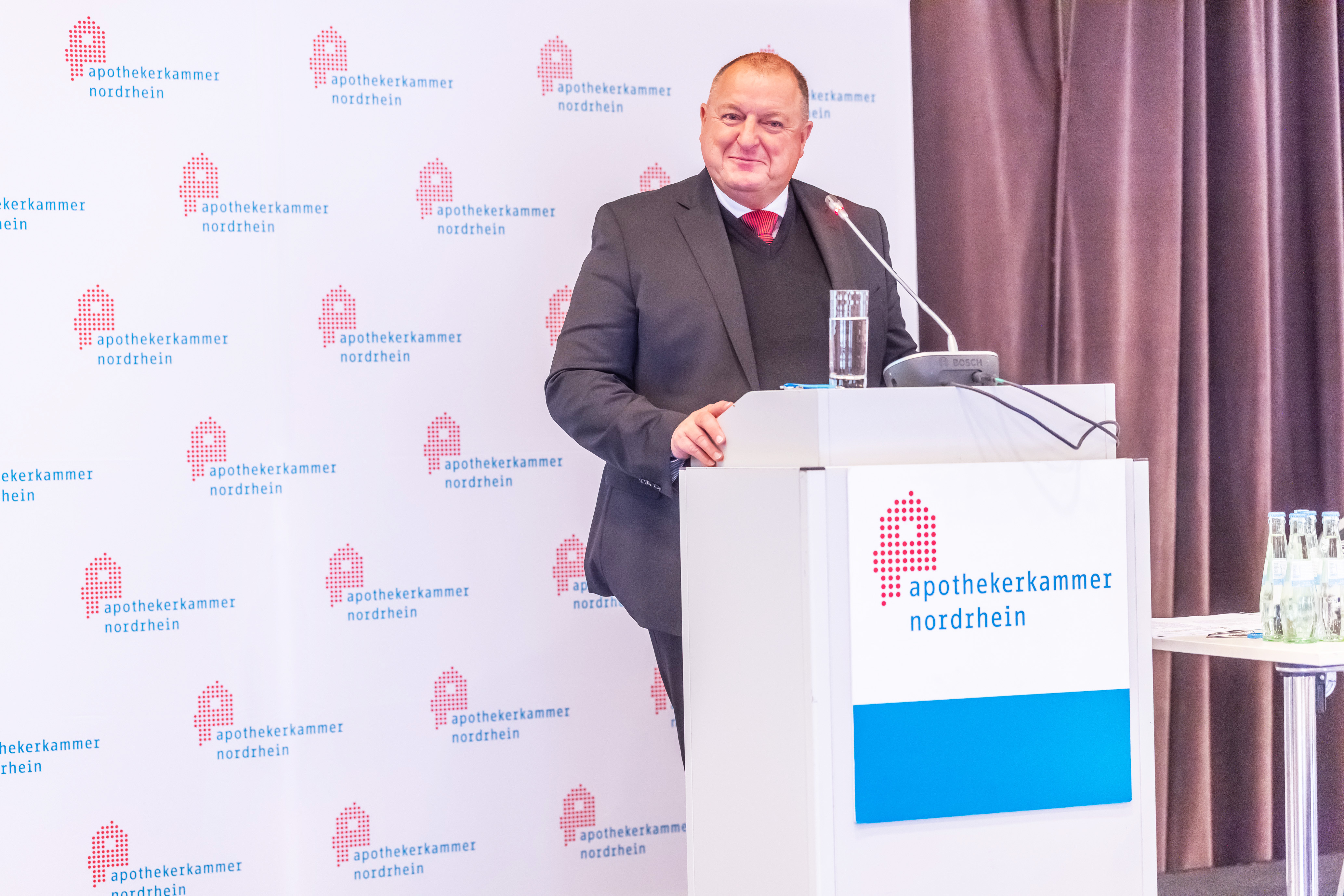 Redner steht an einem Podium mit Mikrofon bei einer Veranstaltung der Apothekerkammer Nordrhein und spricht vor einem Hintergrund mit wiederholtem Kammer-Logo.