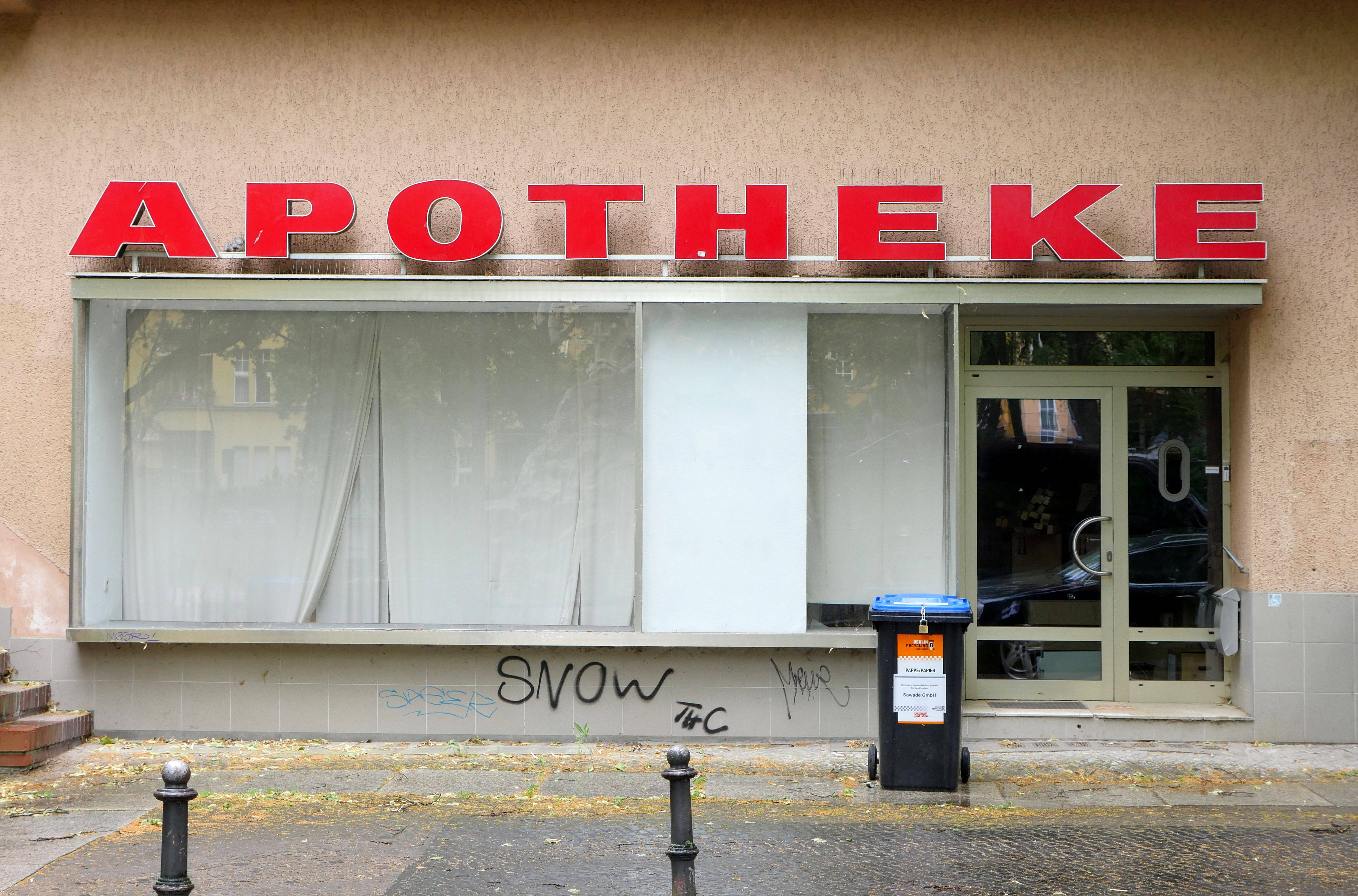 Außenansicht einer geschlossenen Apotheke mit großem roten Schriftzug ‚APOTHEKE‘ über dem Schaufenster. Die Fenster sind mit weißen Vorhängen verhängt, die Fassade ist beige. Unter dem Schaufenster sind Graffiti zu sehen, davor steht eine blaue Mülltonne für Altpapier auf dem Gehweg.