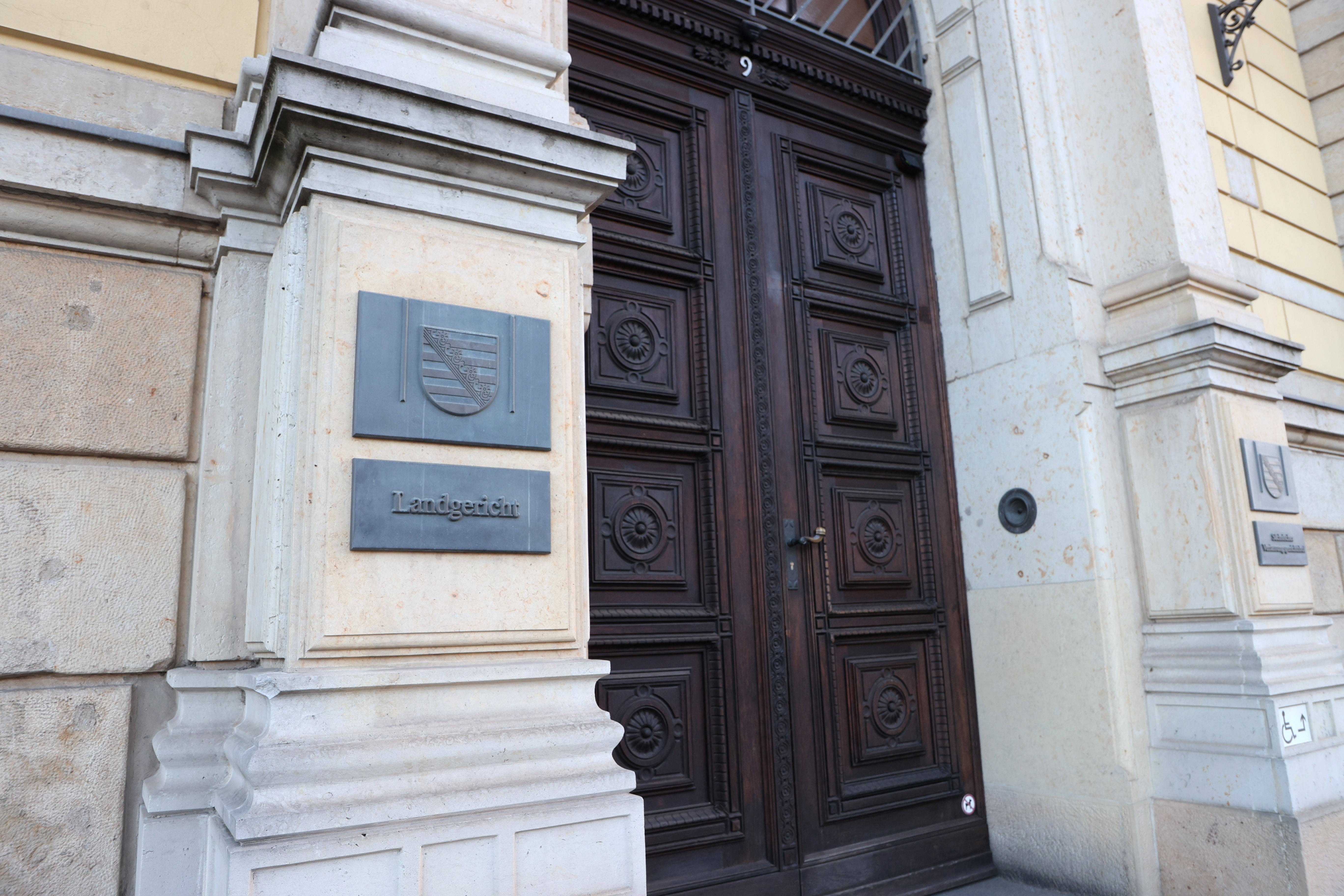 Eingang des Landgerichts Leipzig mit hellen Sandsteinwänden, einem Metallschild mit Wappen und der Aufschrift ‚Landgericht‘ sowie einer großen, dunkelbraunen, verzierten Holztür.