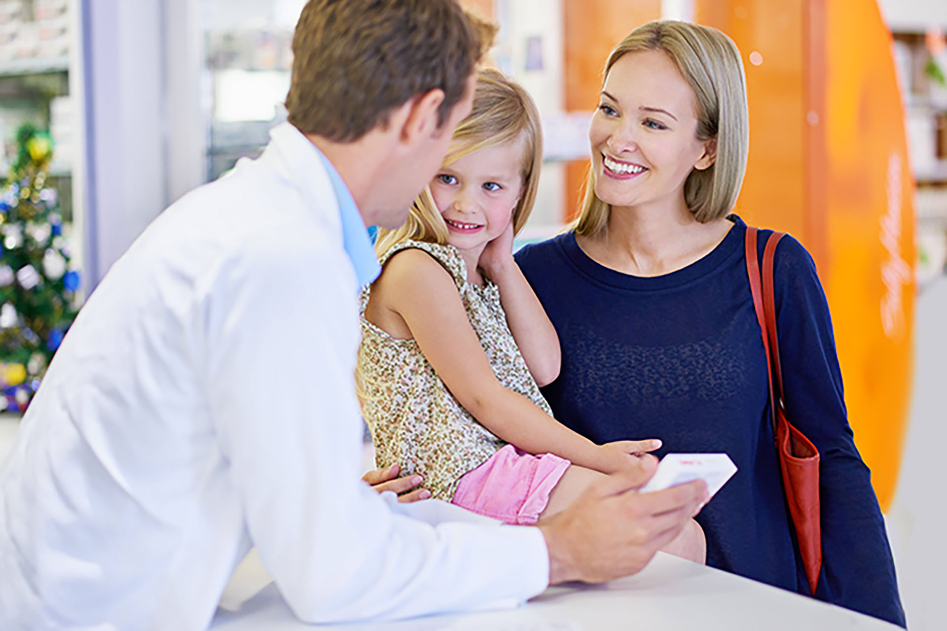 Apotheker im weißen Kittel berät eine erwachsene Person, die ein Kind neben sich sitzen hat. Die drei befinden sich am Handverkaufstisch einer Apotheke, während der Apotheker ein Medikamentenpaket zeigt.