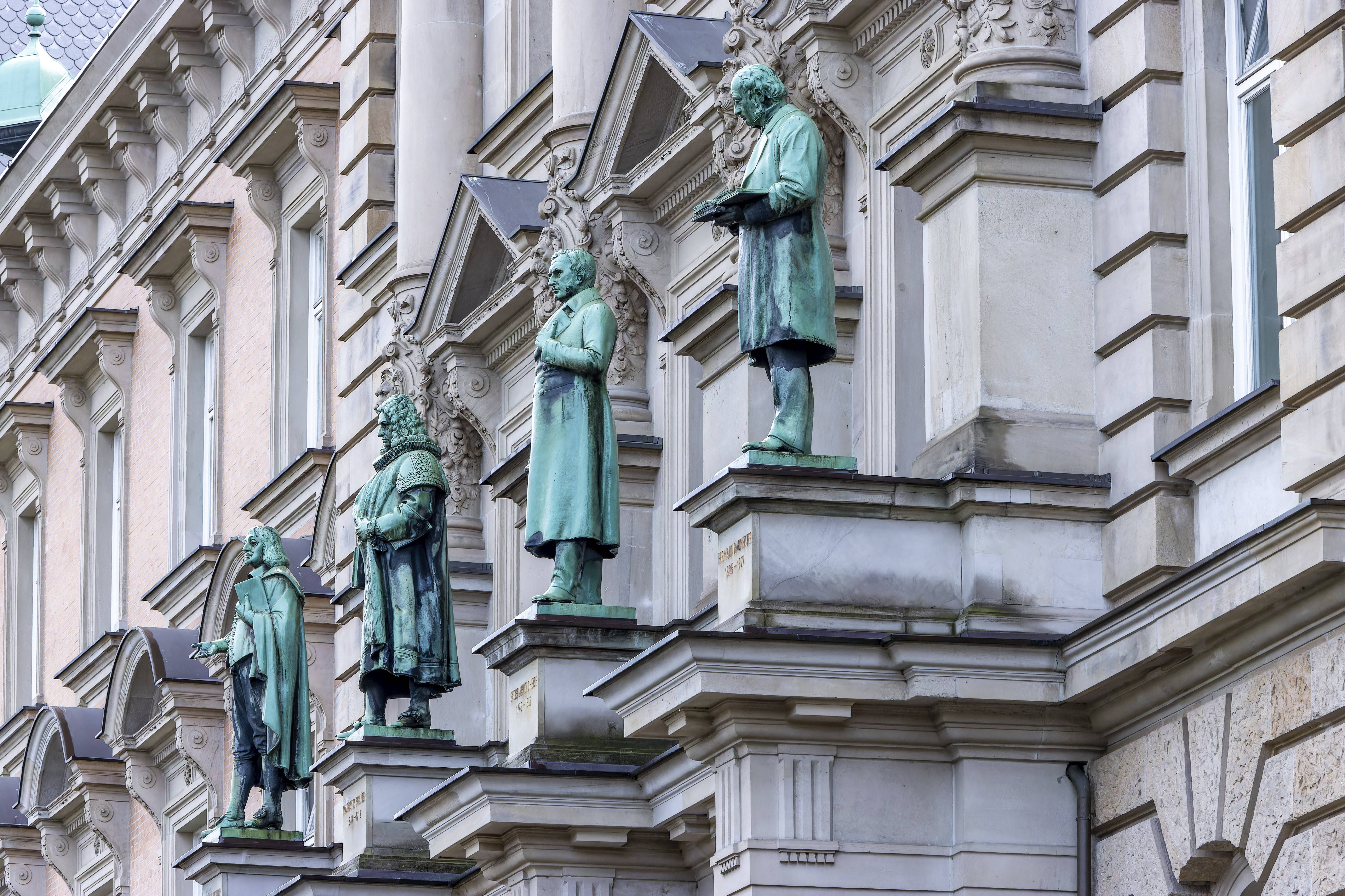 Reihe historischer Statuen aus grünlicher Bronze an der Fassade eines reich verzierten klassischen Gebäudes, jede Figur in unterschiedlicher Pose auf Vorsprüngen oberhalb der Fenster.