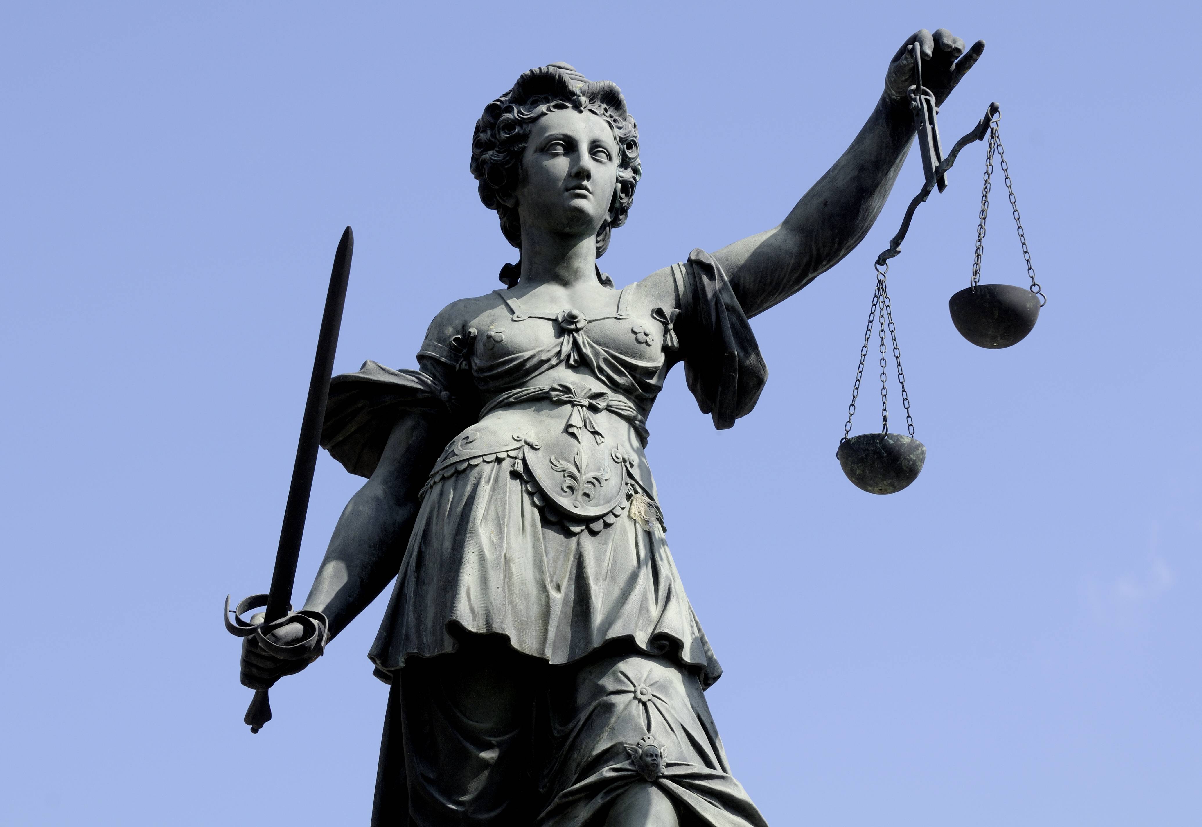 Statue der Justitia vor blauem Himmel, dargestellt mit Schwert in der linken Hand und einer Waage in der rechten Hand, als Symbol für Gerechtigkeit und Rechtsprechung.