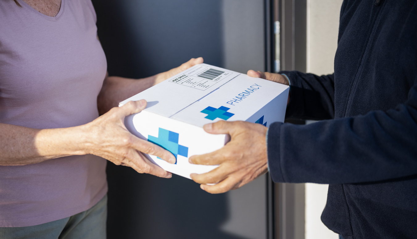 Ein Paket mit blauen Kreuzsymbolen und der Aufschrift ‚Pharmacy‘ wird an einer Haustür von einer Person an eine andere übergeben. Das Paket hat ein Versandetikett und wird von einer Person in dunkler Kleidung an eine Person in heller Kleidung überreicht.