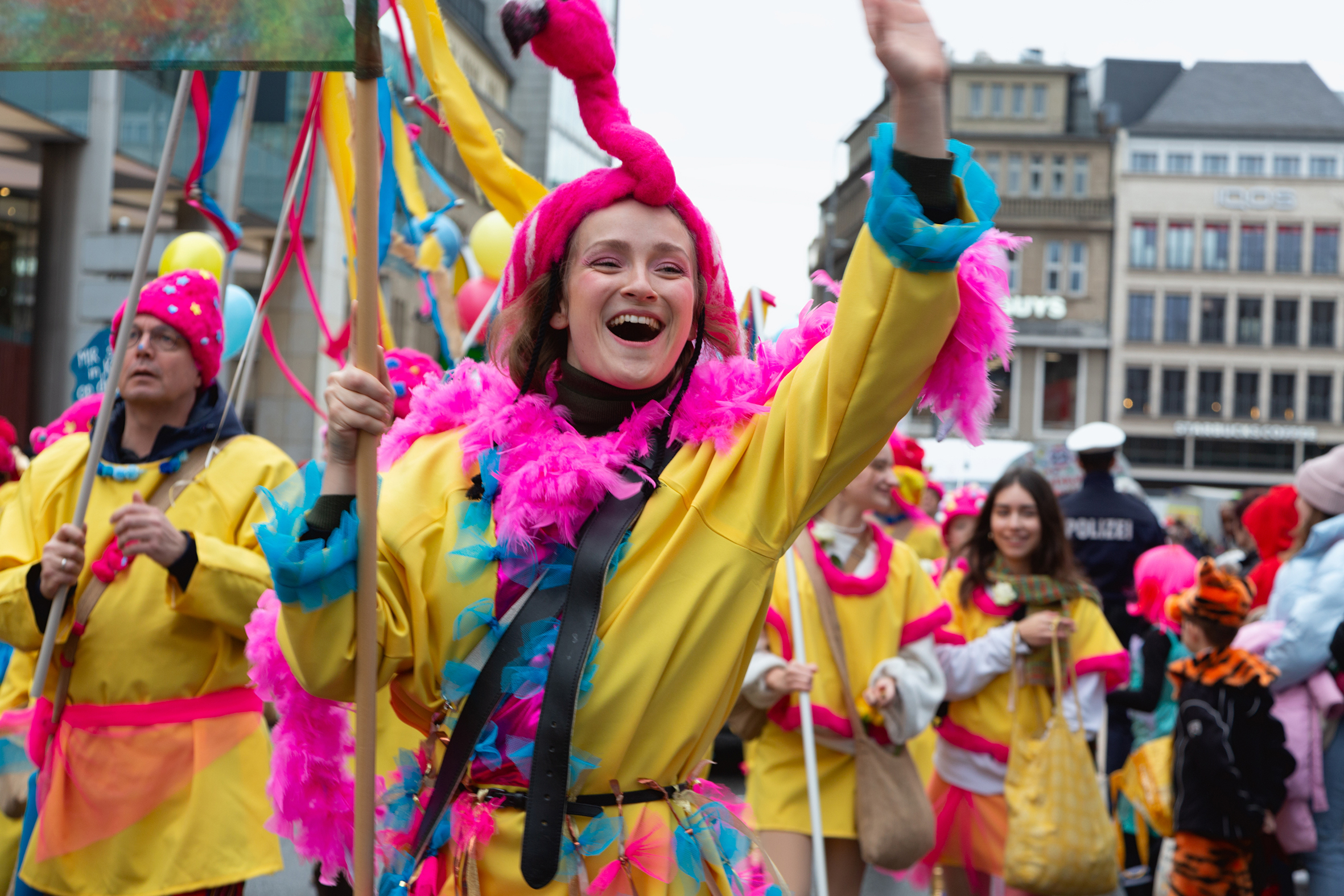 Menschen nehmen an einem farbenfrohen Karnevalsumzug teil. Sie tragen gelbe Kostüme mit pinken und blauen Federn sowie Kopfschmuck in Flamingo‑Form und schwenken bunte Stäbe. Im Hintergrund sind weitere verkleidete Teilnehmende und städtische Gebäude zu sehen.