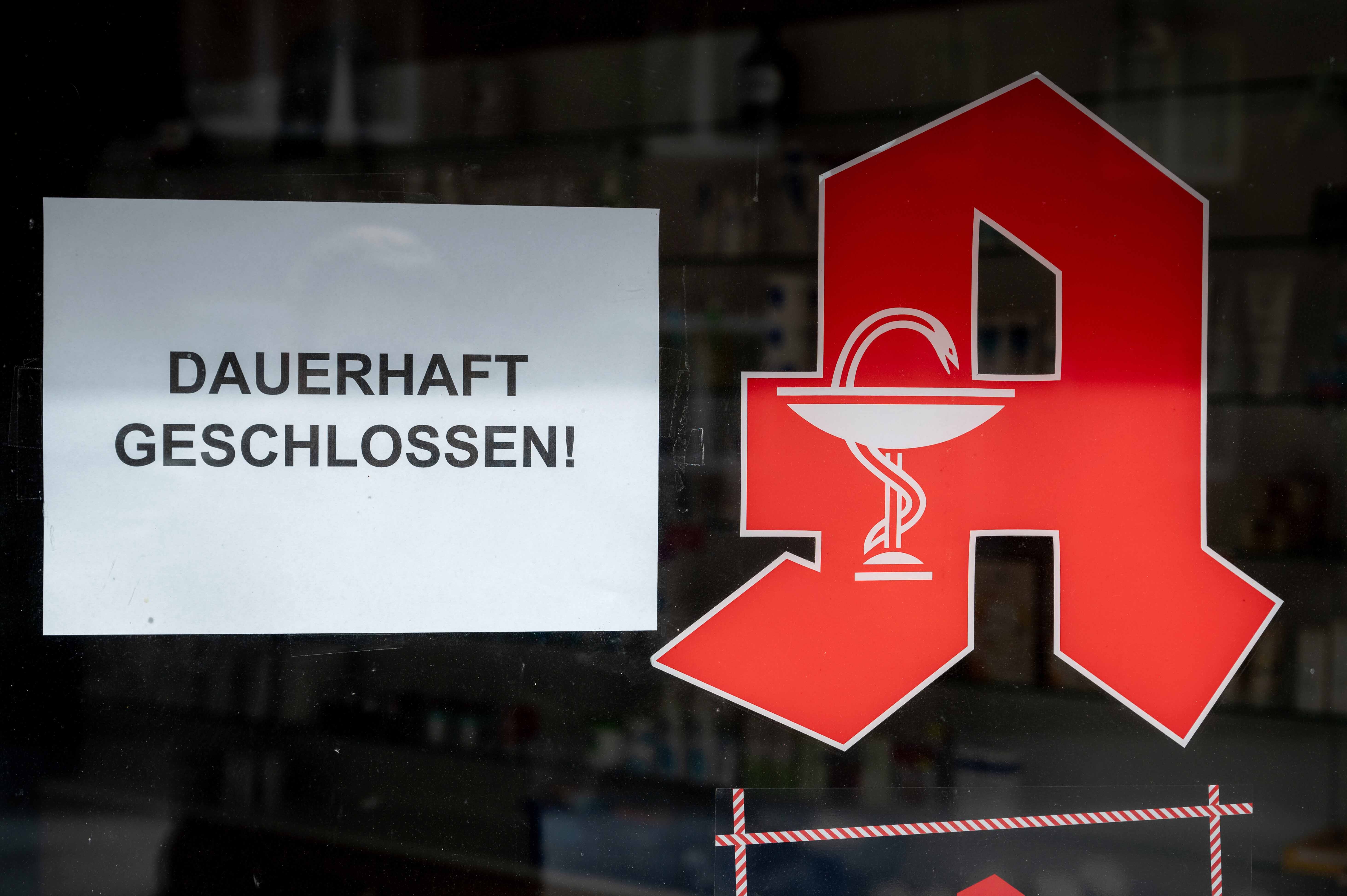 Schaufensterscheibe einer Apotheke mit einem weißen Aushang „DAUERHAFT GESCHLOSSEN!“. Daneben ist ein großes rotes Apotheken‑A mit Schale und Schlange zu sehen; im Hintergrund sind unscharf Regale im Innenraum erkennbar.