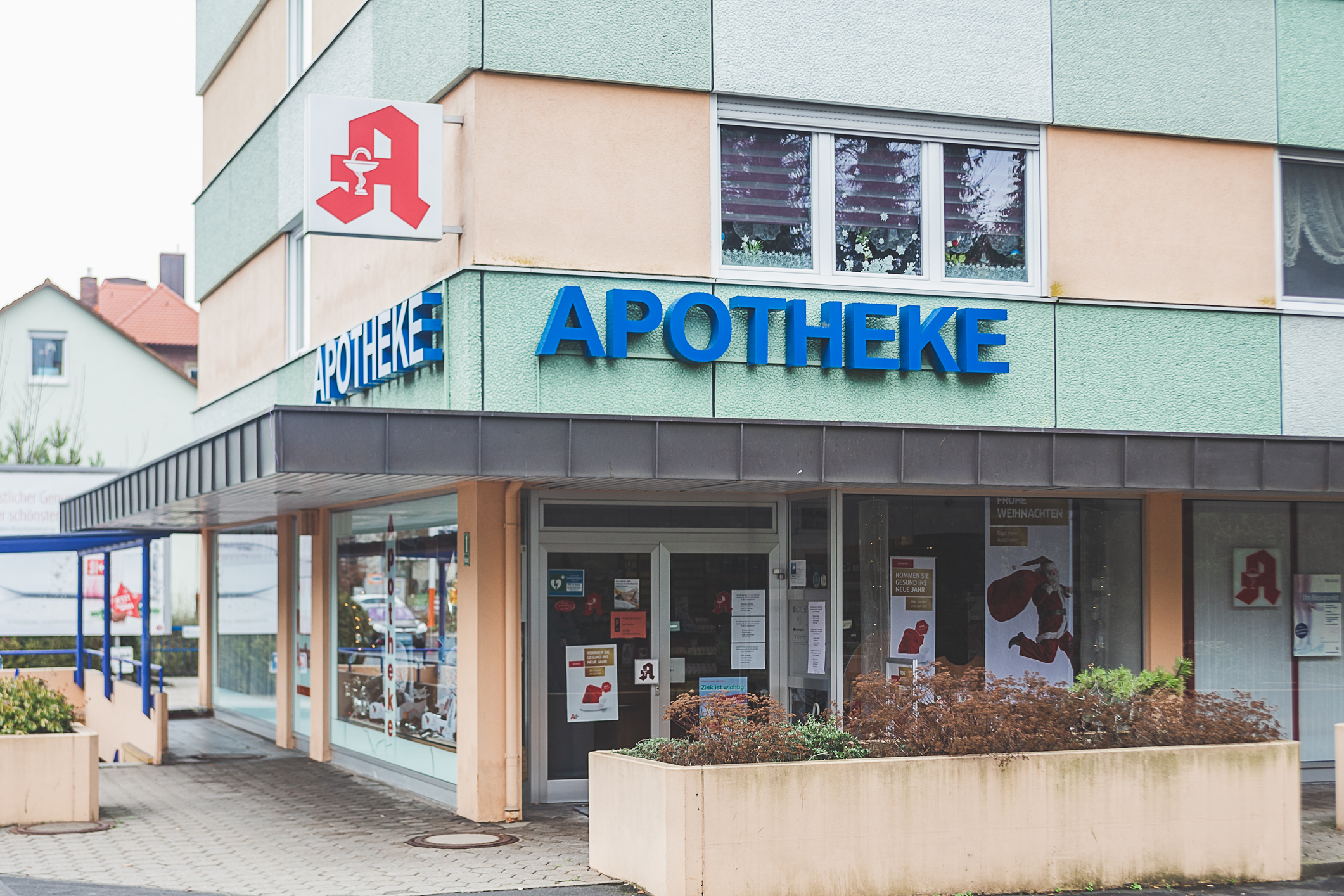 Außenansicht einer Apotheke in einem mehrgeschossigen Gebäude. Über dem Eingangsbereich sind das rote Apotheken-A und der blaue Schriftzug „APOTHEKE“ zu sehen. Vor dem Gebäude befinden sich Schaufenster, Pflanzbeete und ein gepflasterter Gehweg.